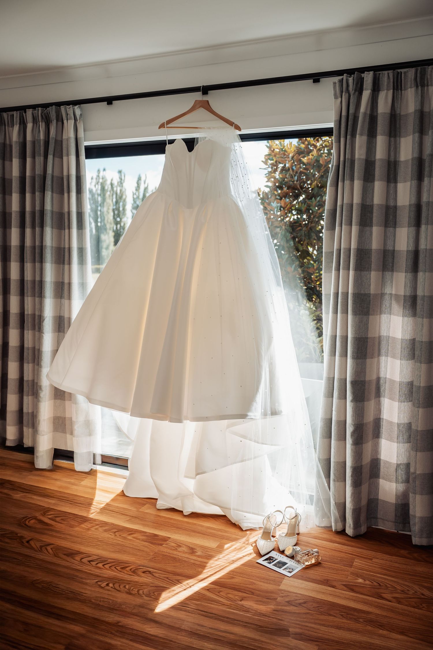 White ballgown wedding dress hanging by window with bridal heels below on hardwood floor.