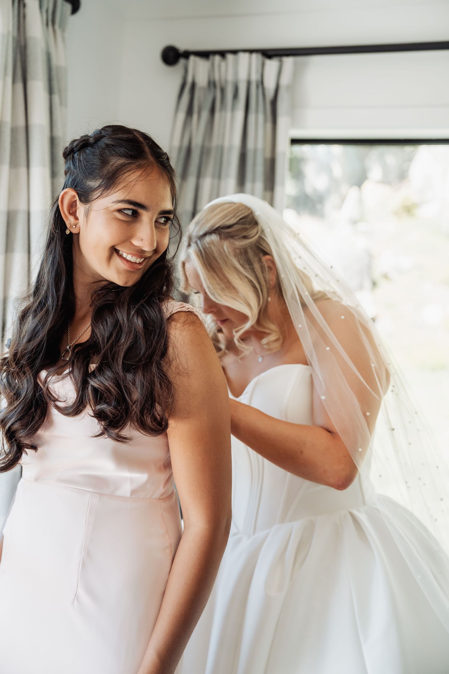 Bridesmaid in blush dress smiling as bride in white gown and veil adjusts her shoulder.