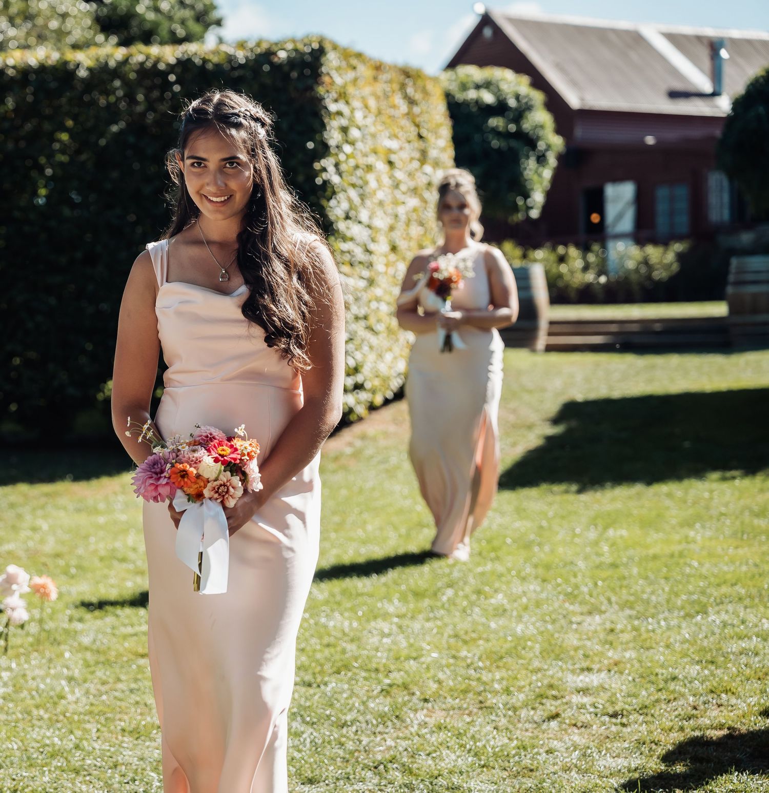 Bridesmaid in blush satin gown holding colorful bouquet walks down outdoor wedding aisle on sunny day.