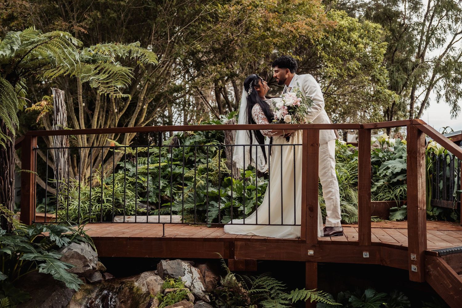 Bride and groom sharing a kiss on a wooden bridge surrounded by lush tropical garden greenery.