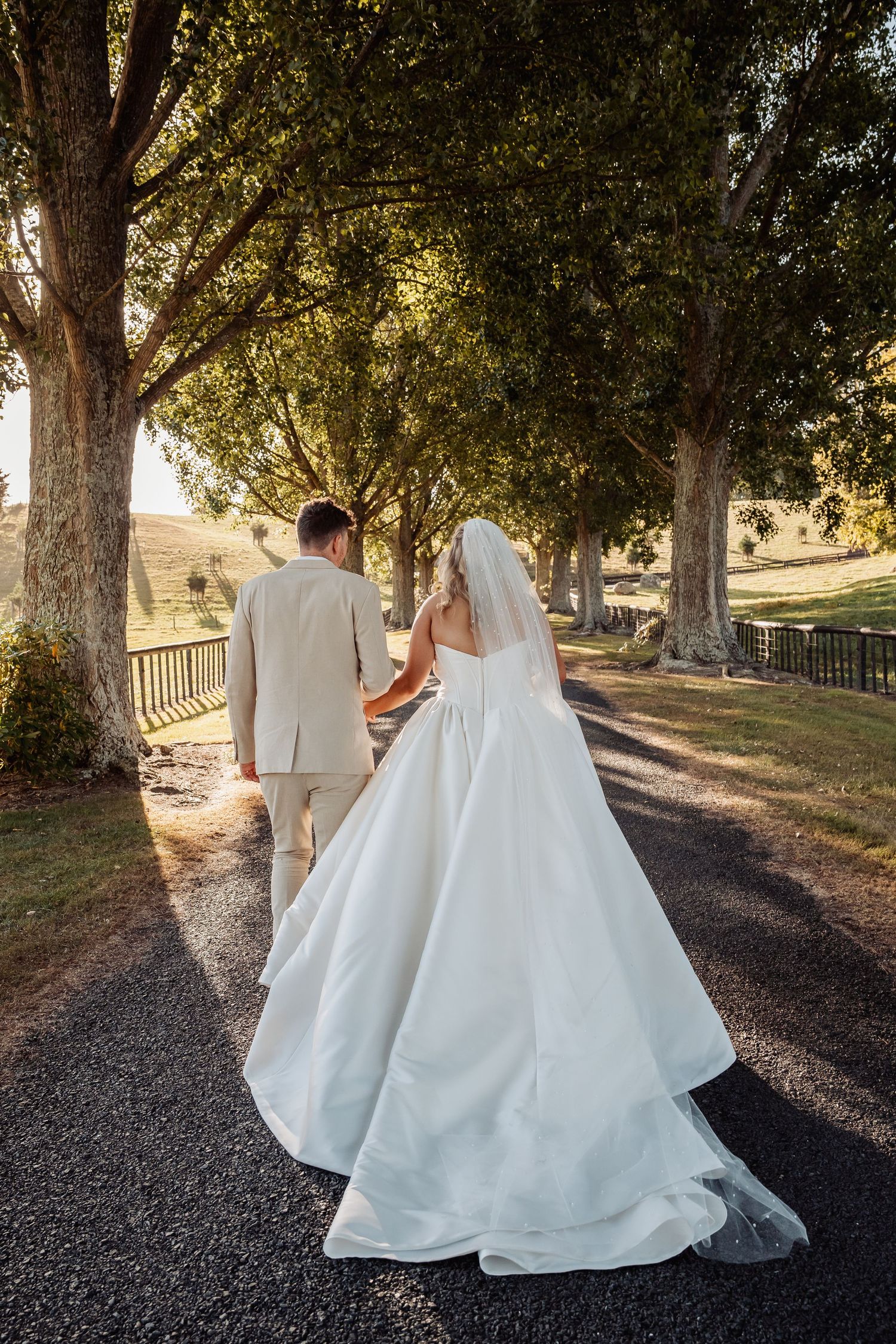 Bride and groom walking hand in hand down a tree-lined path, bride in white gown with veil and train.