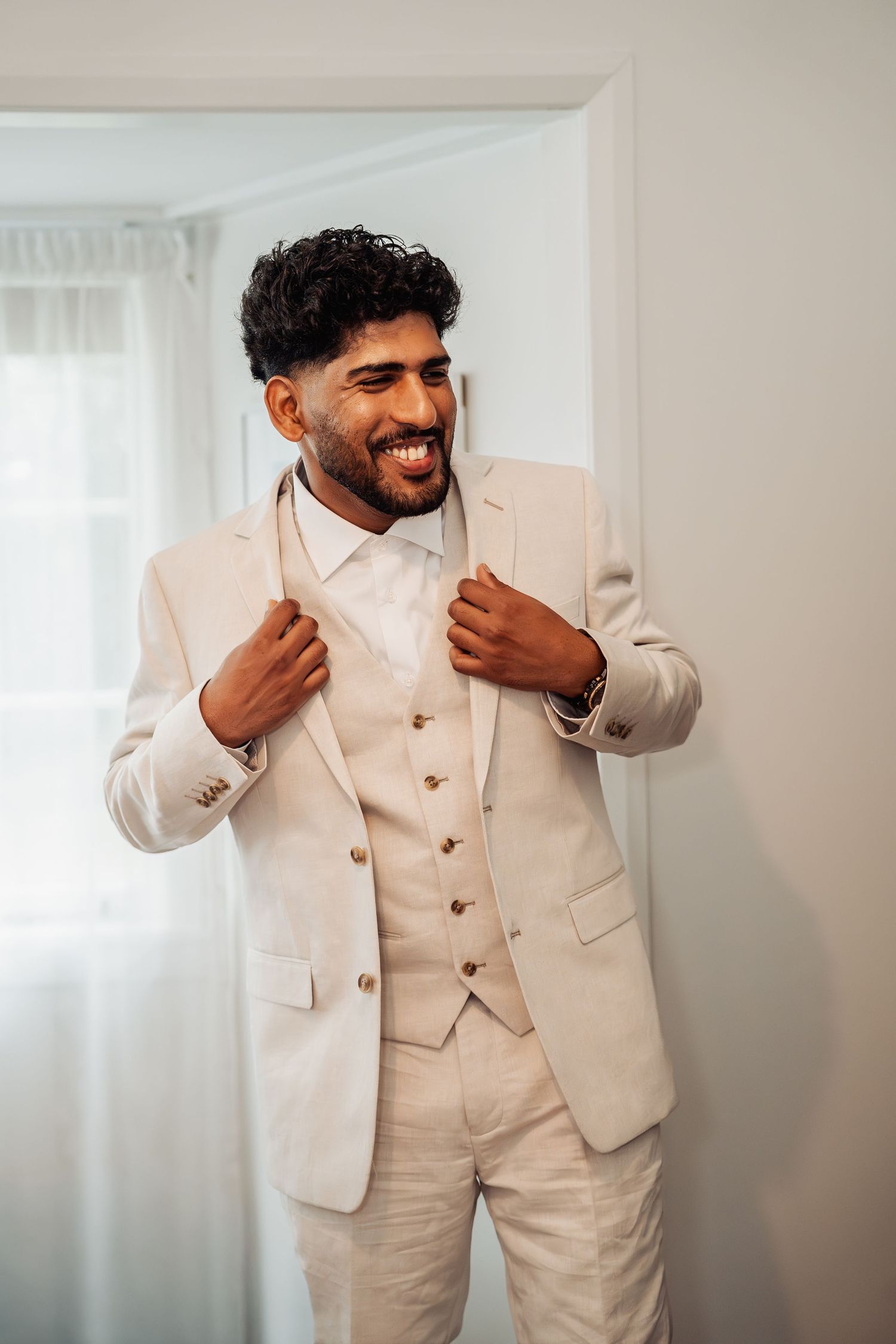 Smiling groom in elegant cream three-piece suit adjusting jacket lapels while standing by a bright window.