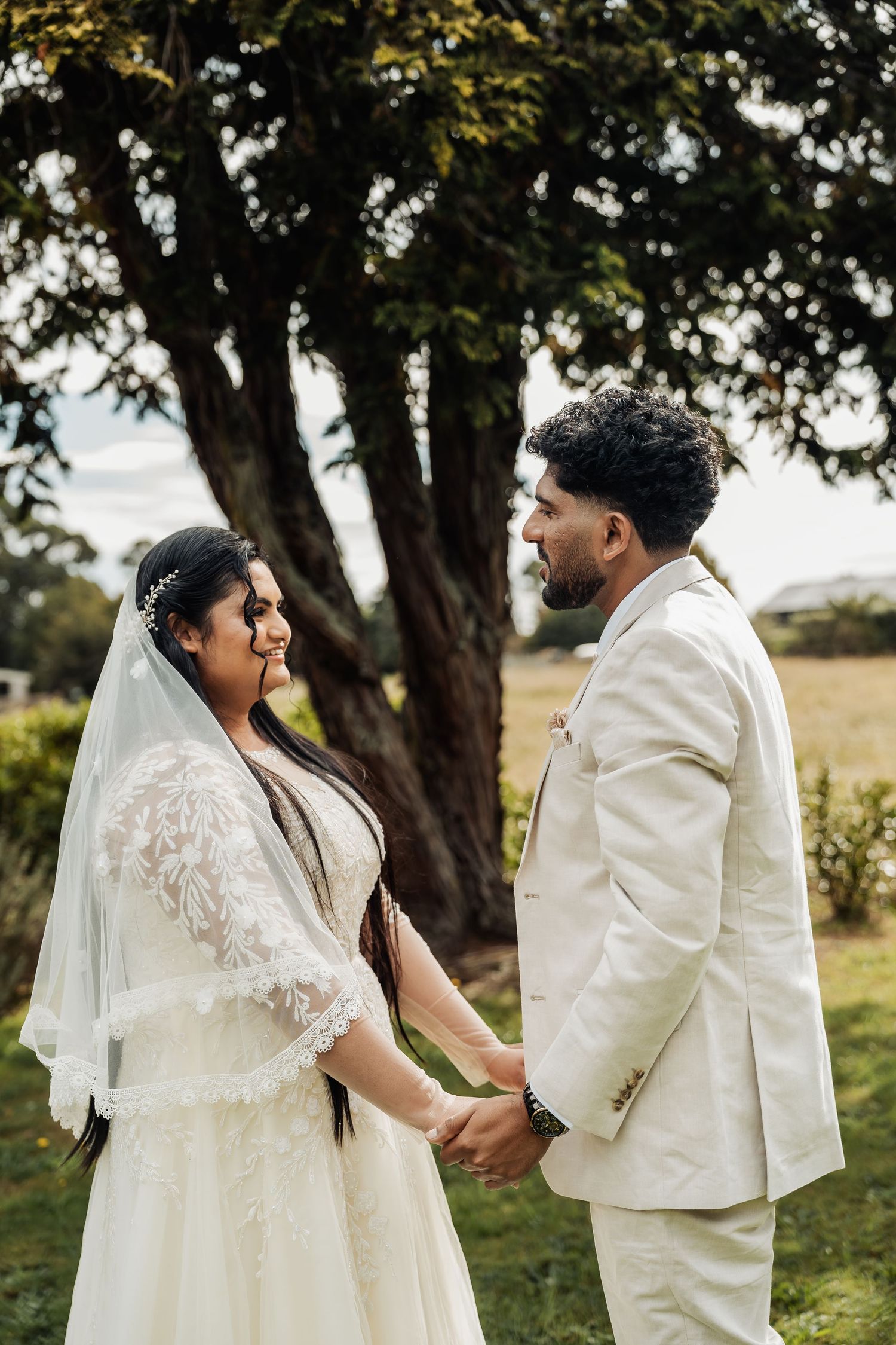 Bride and groom holding hands and gazing at each other outdoors under a large tree on wedding day.