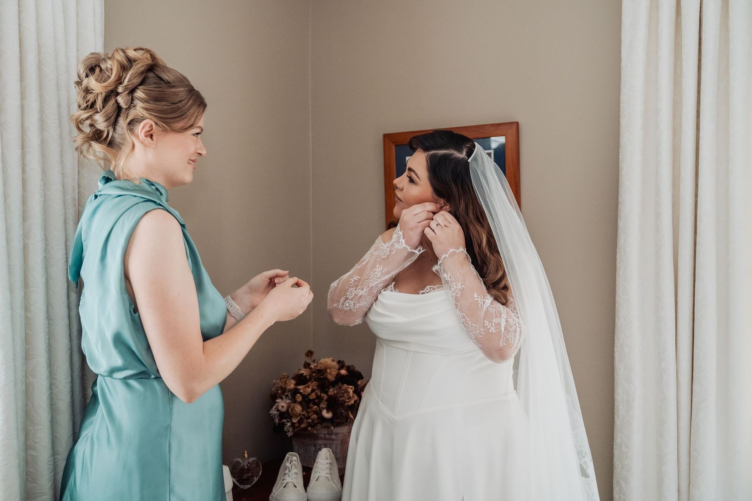 Two people prepare in a bridal suite on wedding day, with a woman in mint green helping adjust a white gown.
