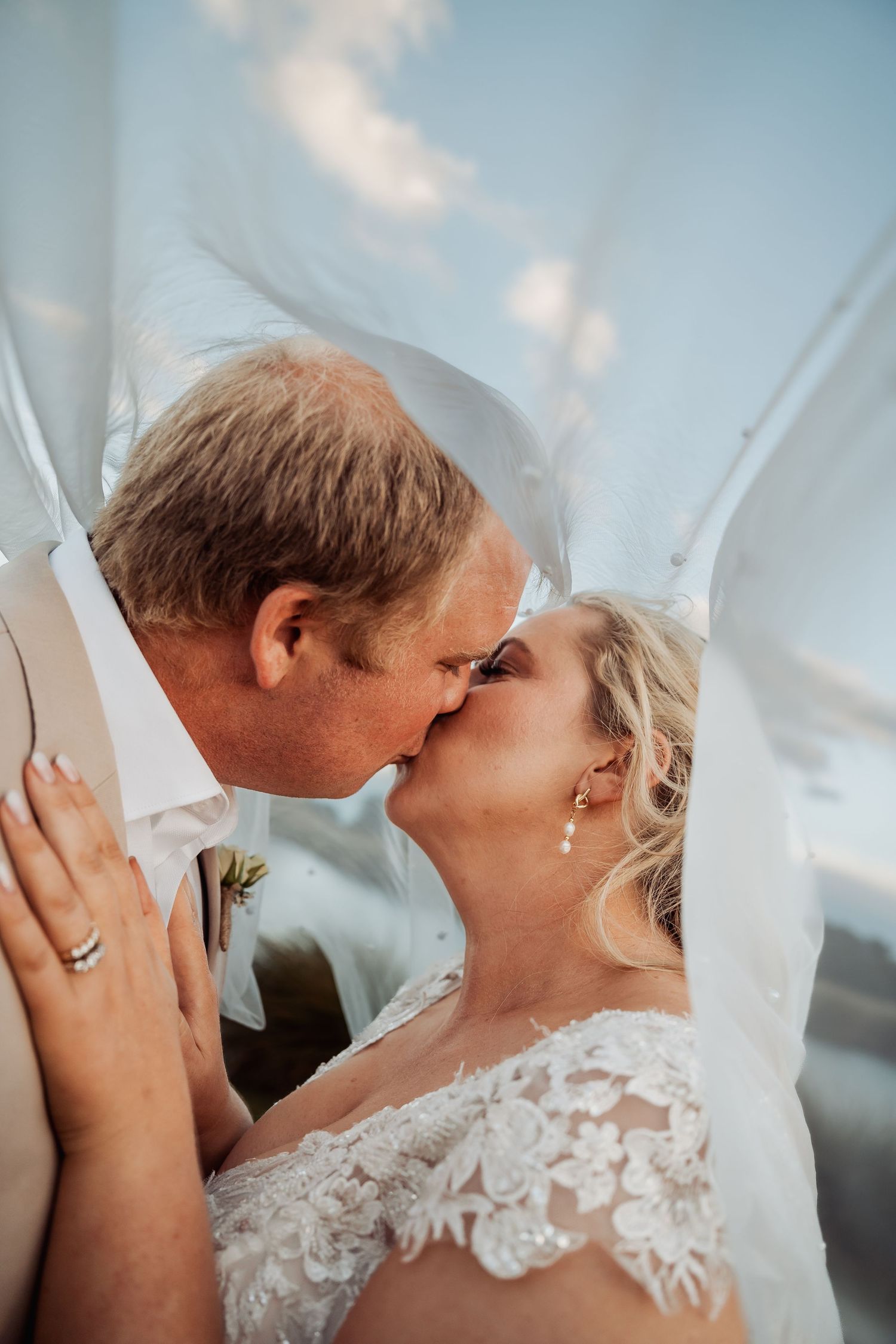Newlyweds share an intimate kiss under the bride's veil with blue sky background.