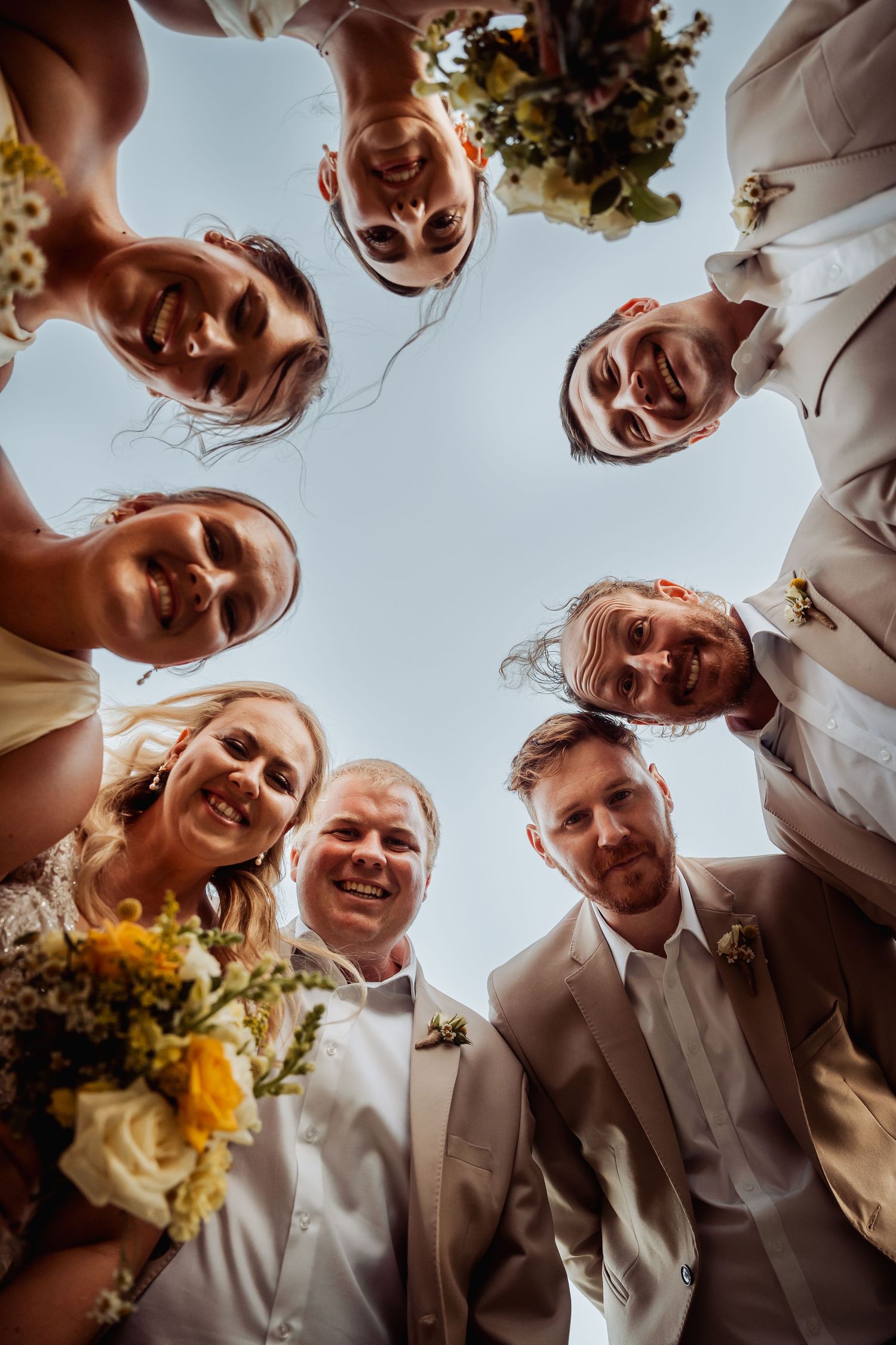 Wedding party huddles together smiling in circle formation photographed from below against blue sky.