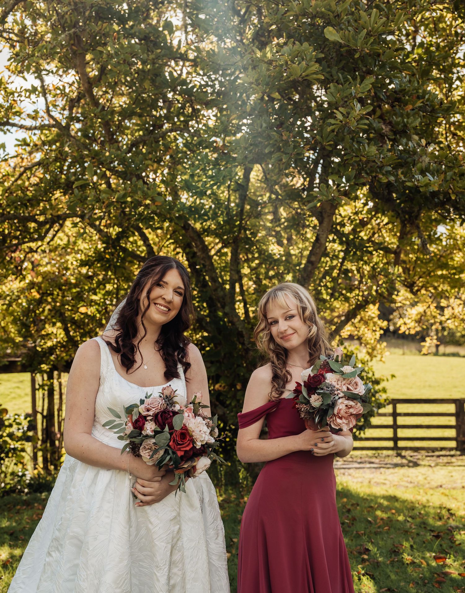 Bride in white lace gown and bridesmaid in burgundy dress holding bouquets outdoors.