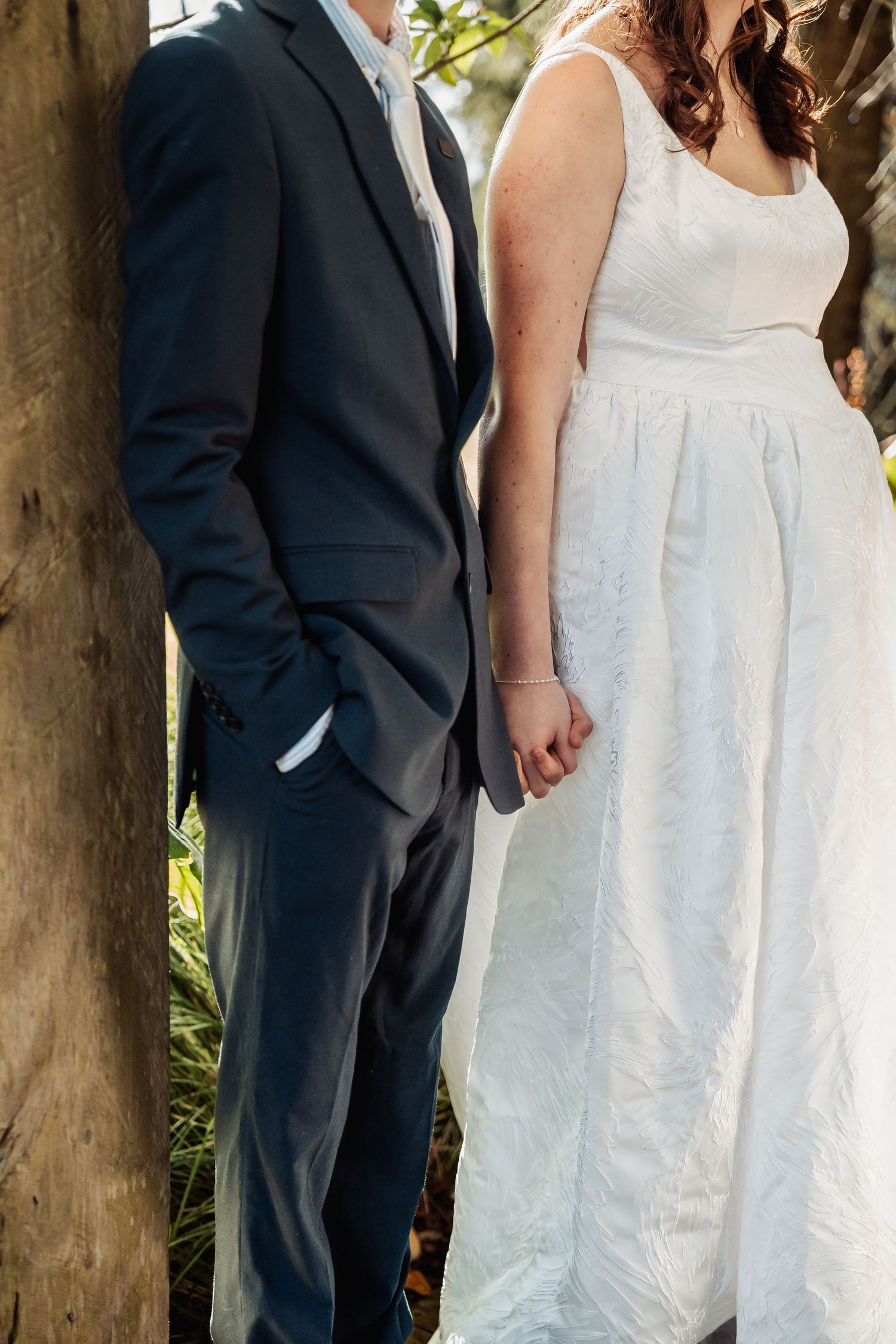 Bride and groom in white dress and dark suit hold hands beside a tree outdoors.