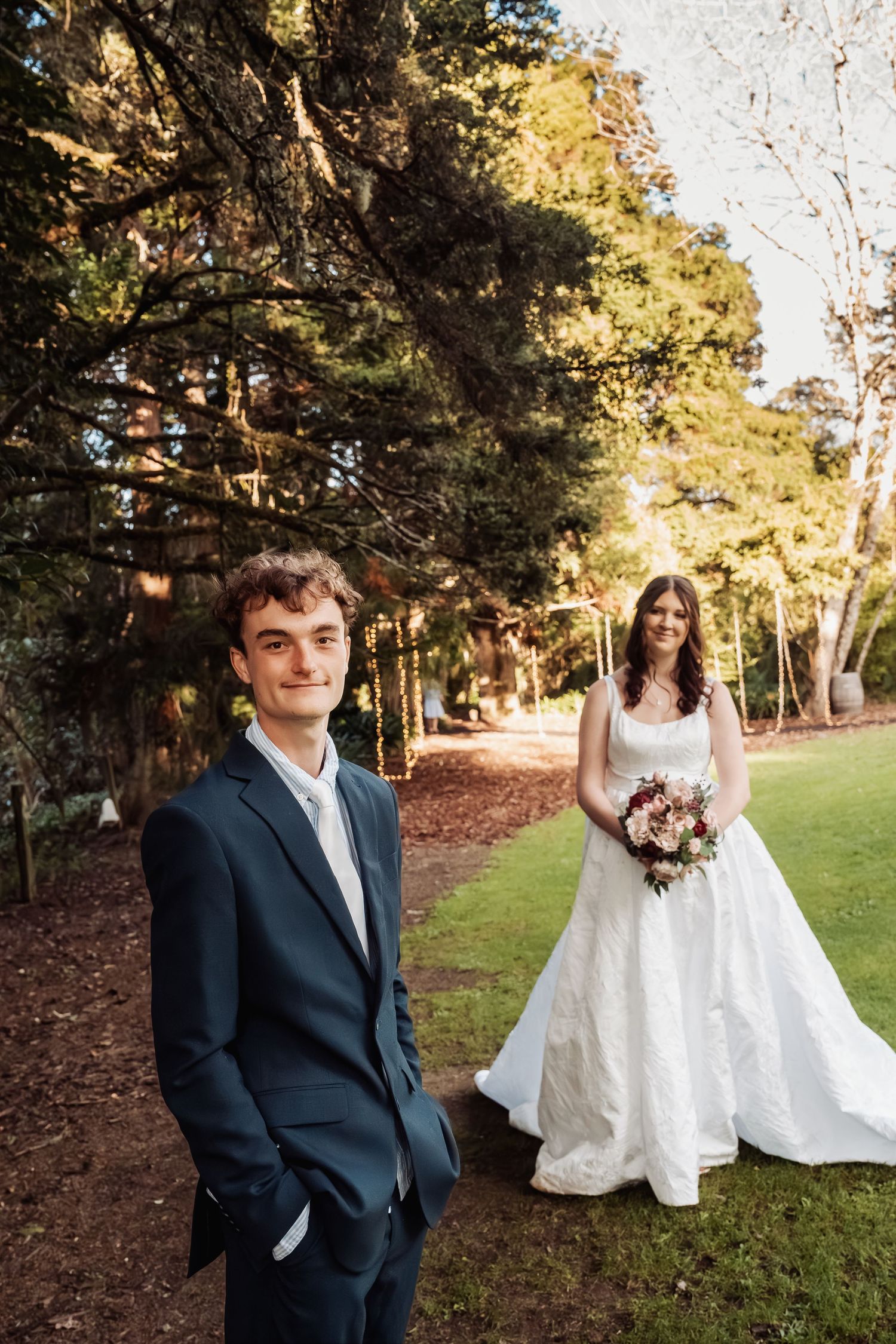 Groom in navy suit stands before bride holding bouquet under tall trees at wedding.