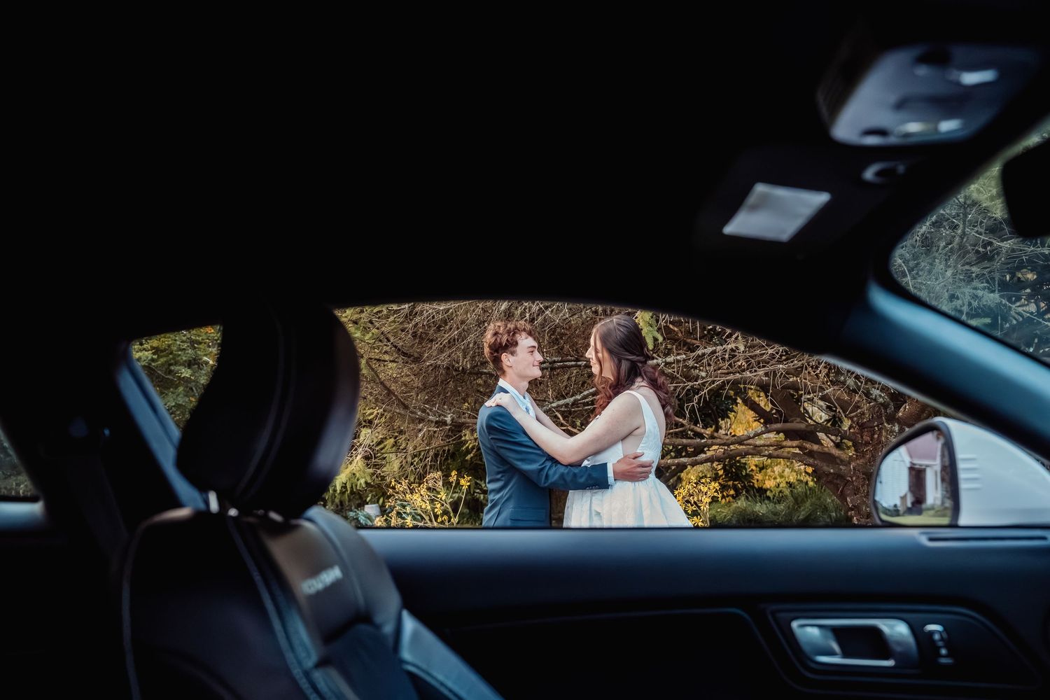 Bride and groom embrace outdoors, framed beautifully through the window of a white sports car in autumn setting.
