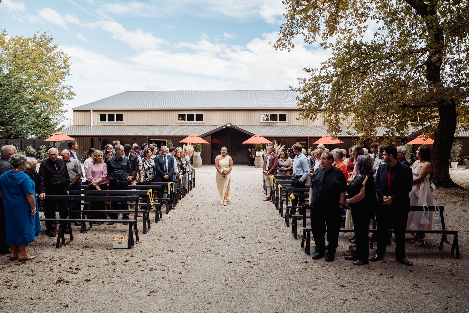 Outdoor wedding ceremony with guests seated on wooden benches at rustic barn venue.