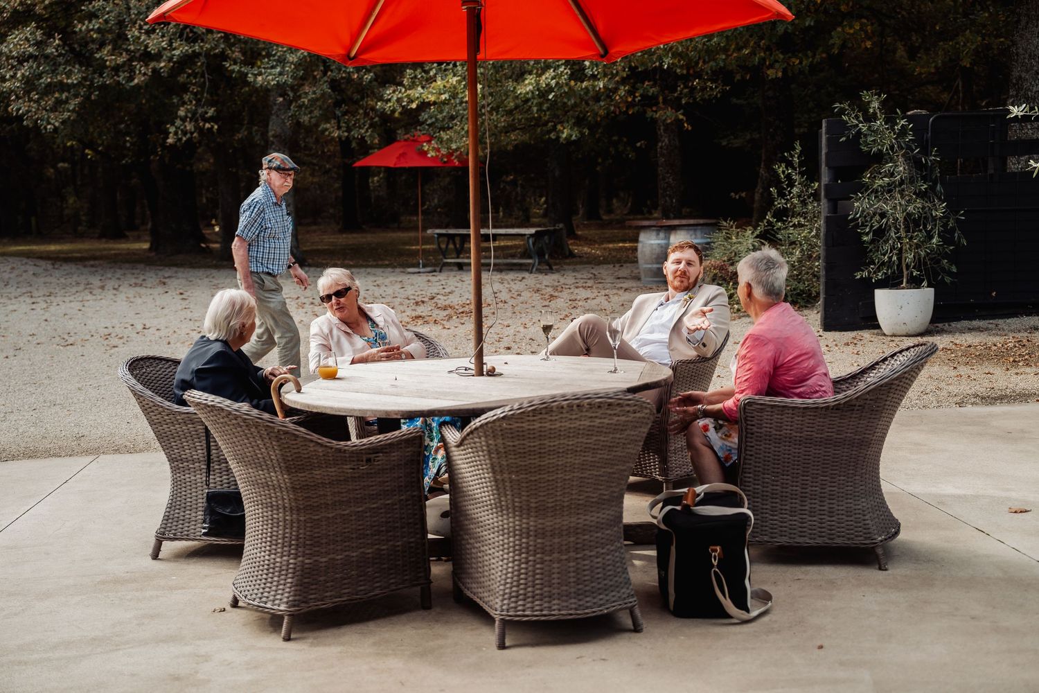 Group of guests seated around a round wicker table under a red patio umbrella outdoors.