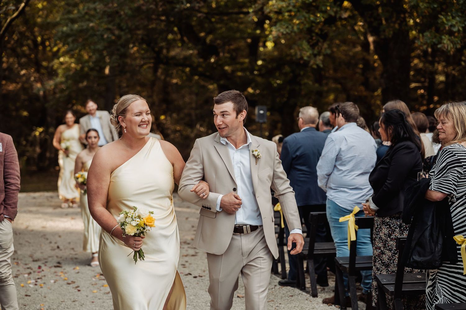 Bride in yellow dress walks down sandy aisle with groom in beige suit at outdoor wedding.