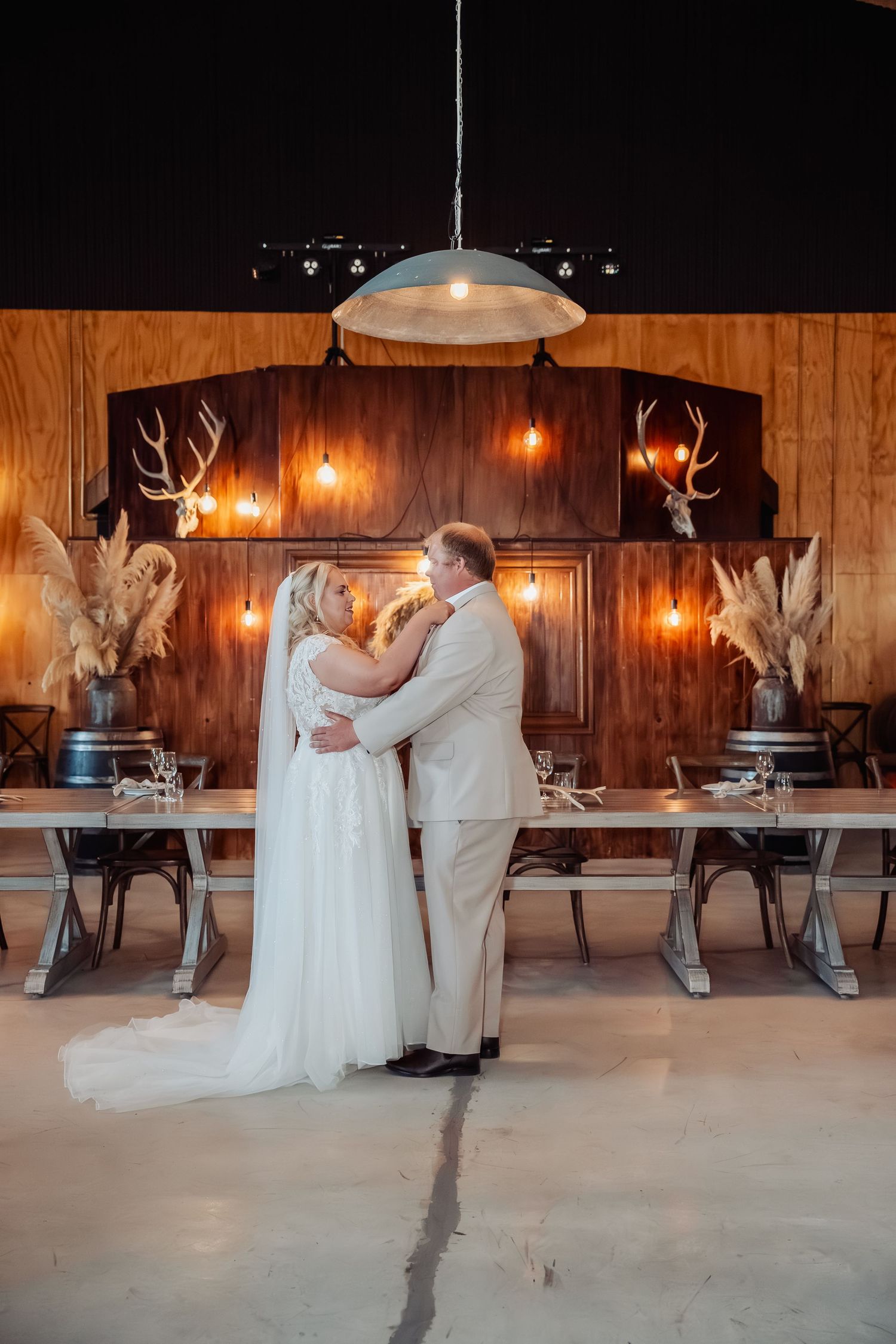 Bride and groom share romantic embrace in rustic barn venue with warm Edison bulb lighting.