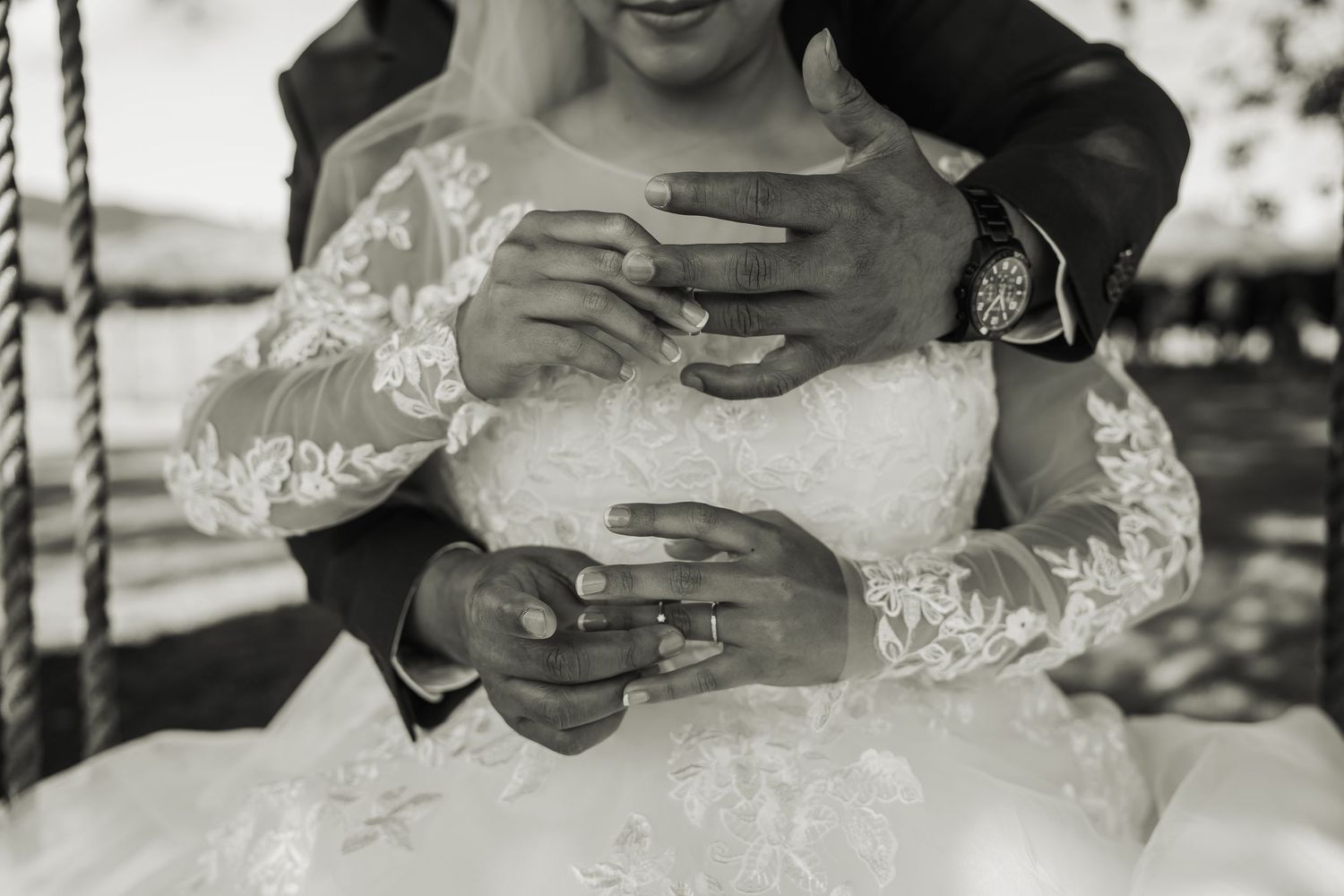Black and white close-up of bride and groom showing wedding rings while embracing on swing.