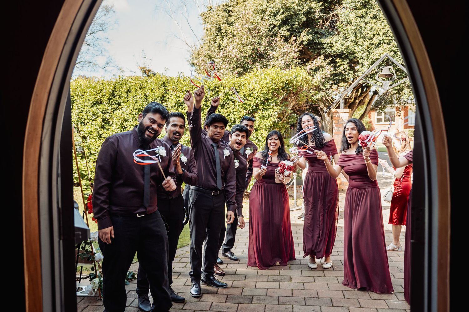 Wedding party in burgundy attire posing with props at venue entrance.