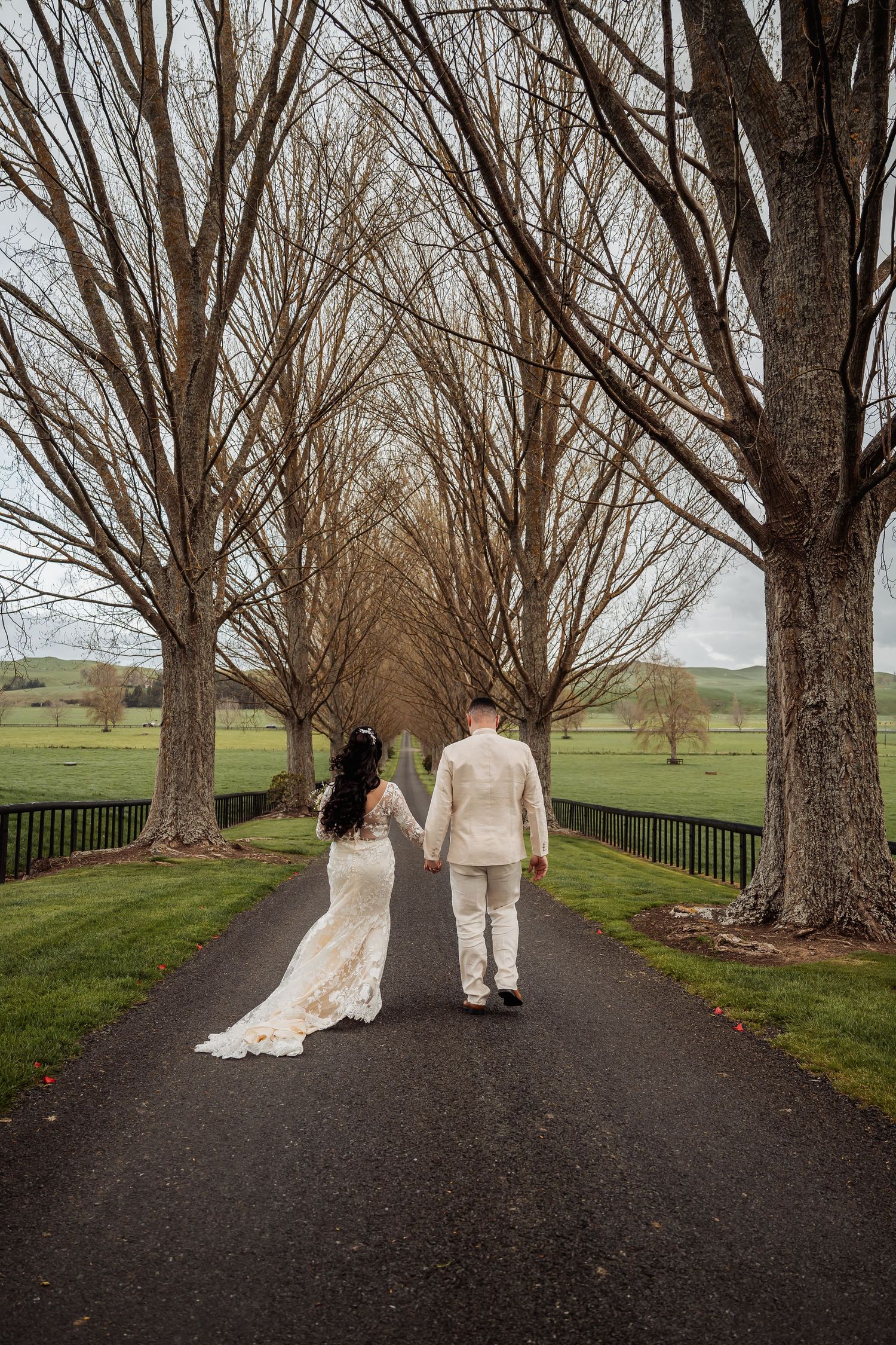 Couple walks down a tree-lined path in wedding attire during autumn.