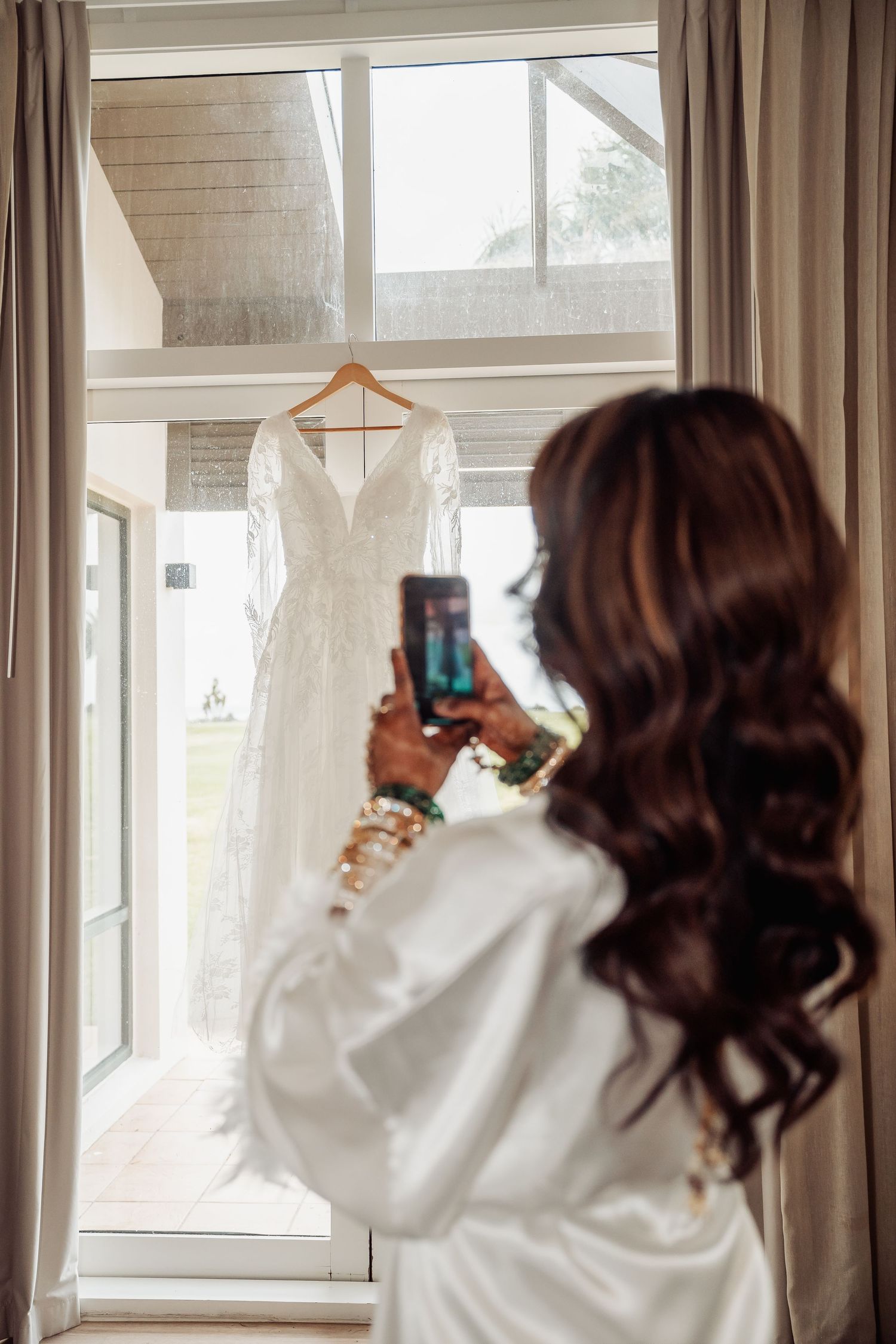 Three bridesmaids in white robes take photos through windows in a bright sunlit room.
