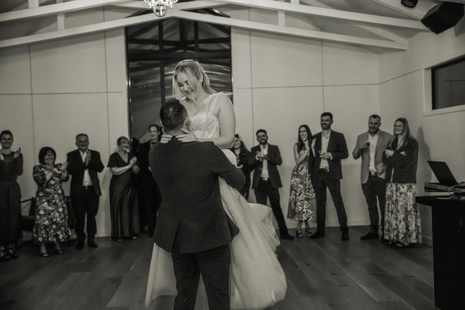 A couple shares their first dance at a wedding reception while guests watch from the edges of a rustic venue hall.