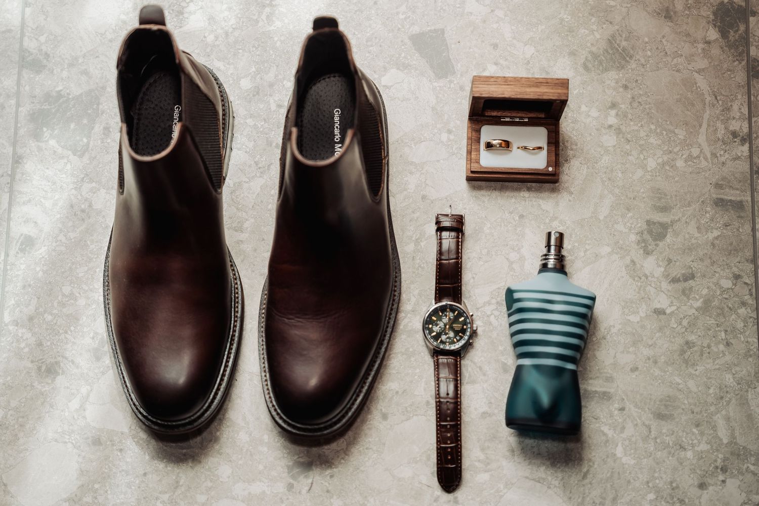Wedding accessories including brown leather shoes, watch, and tie arranged neatly on a light surface.