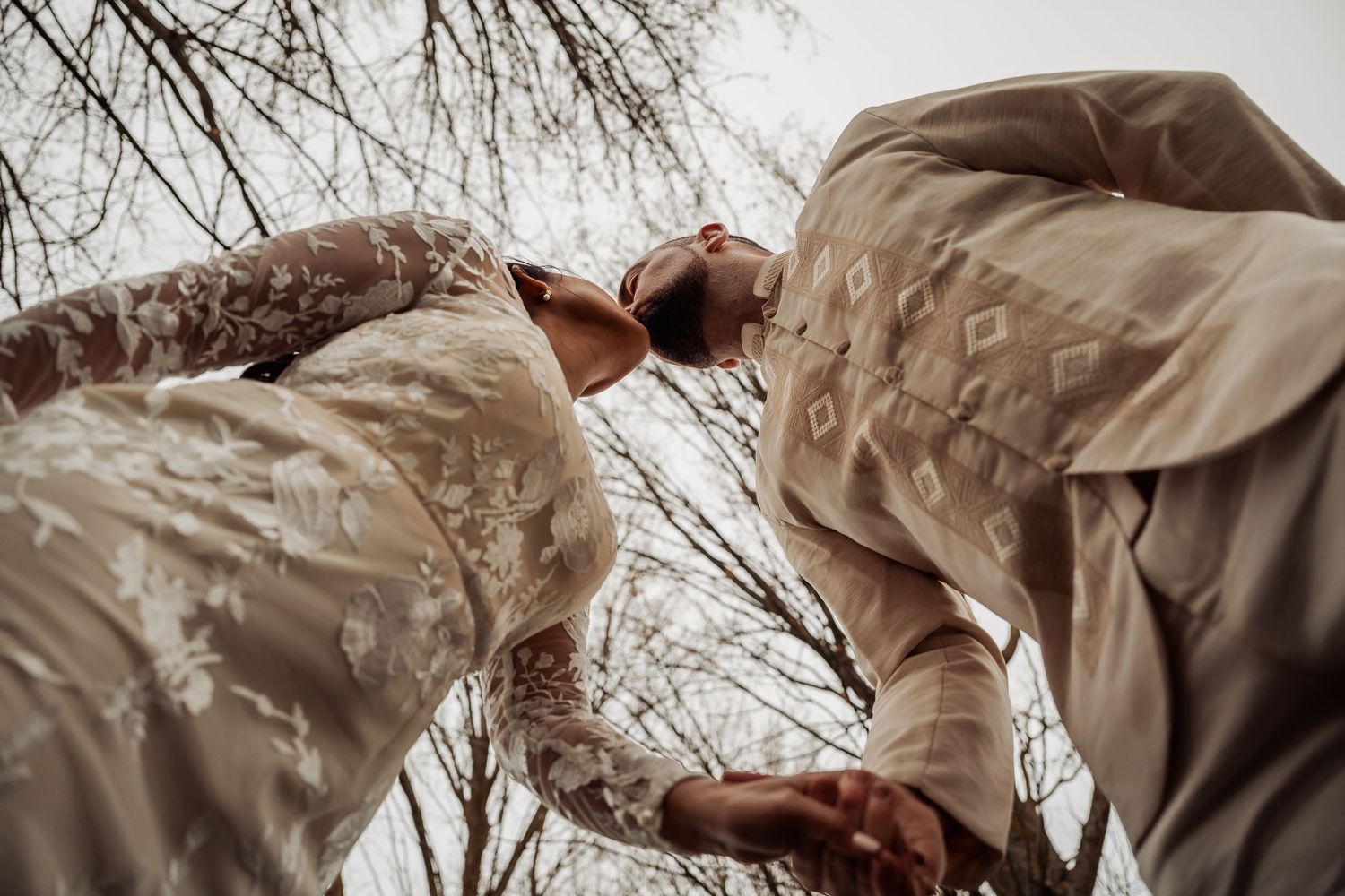 A low-angle view of two people in beige clothing reaching toward each other against a backdrop of bare tree branches.