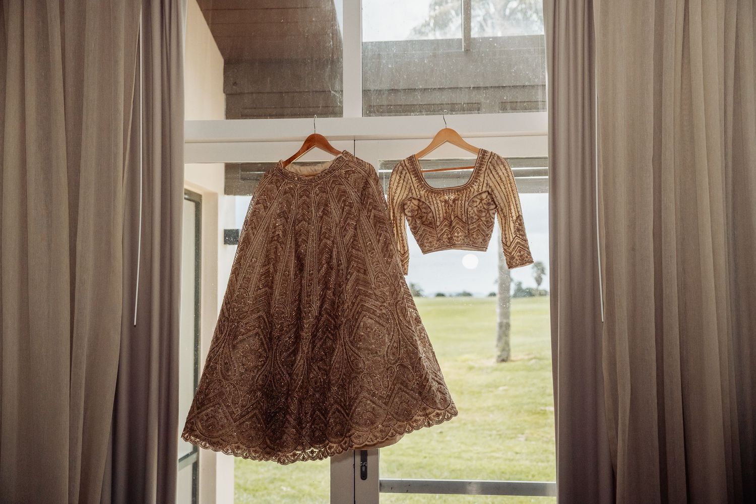 Brown formal dresses hanging on hangers against a window with sheer curtains and natural lighting.