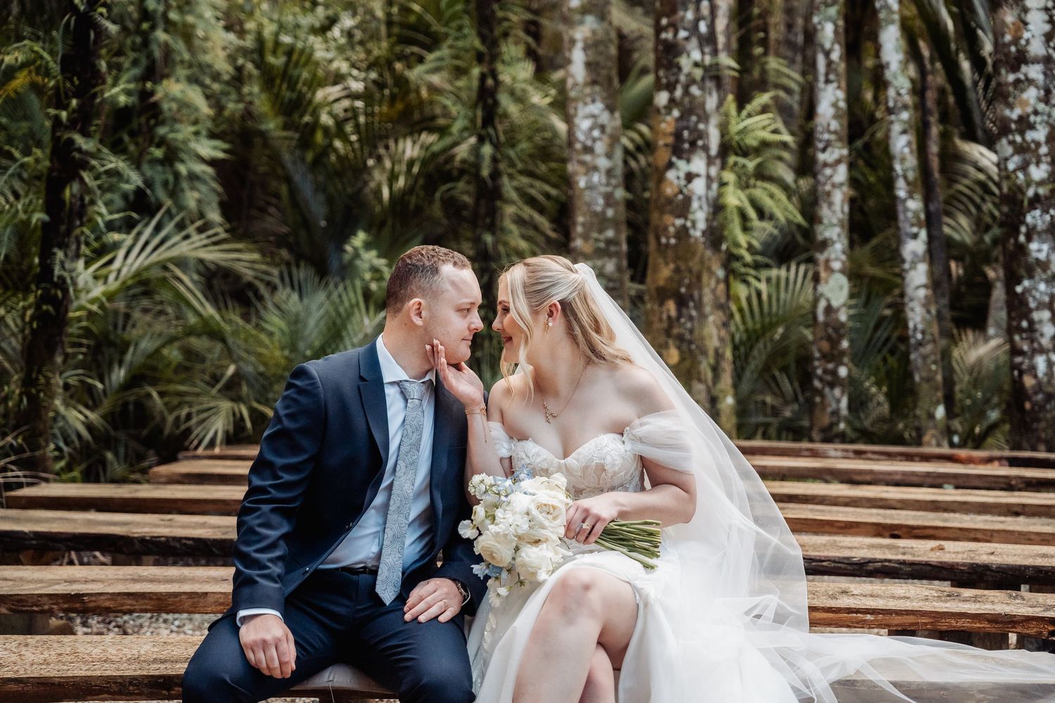 Wedding couple seated on wooden benches in a forest setting with palm trees.