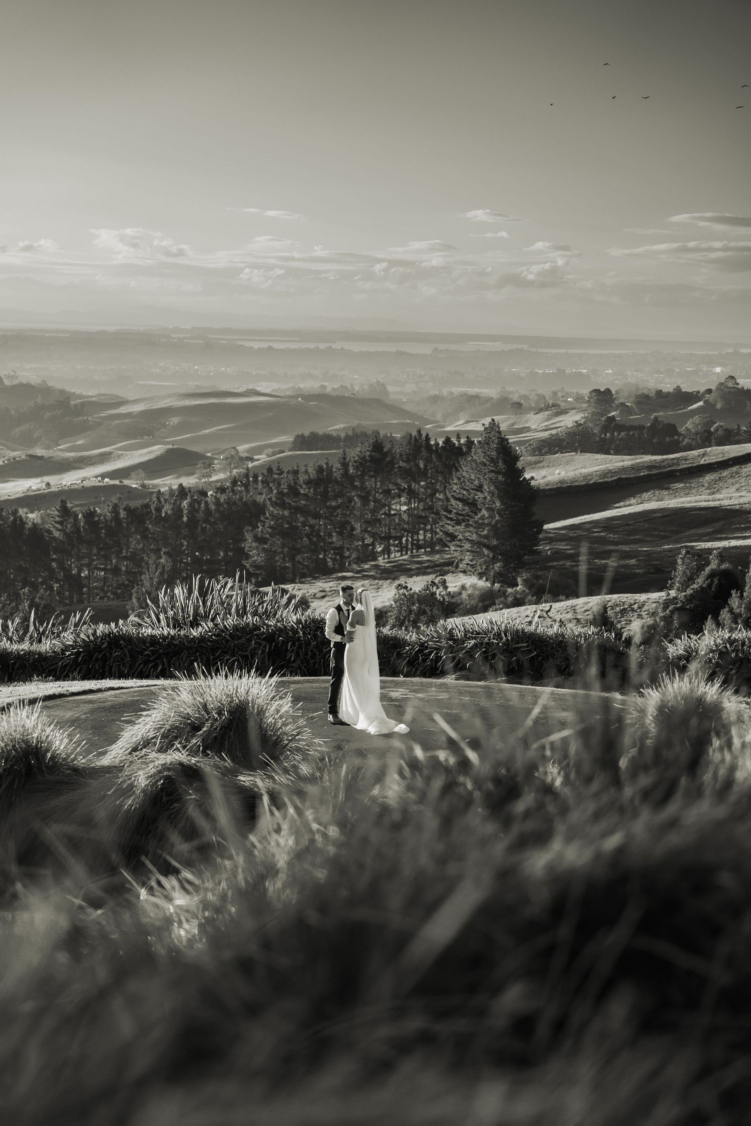 Black and white photo of a bride and groom embracing on a hilltop with rolling countryside and dramatic sky behind them.