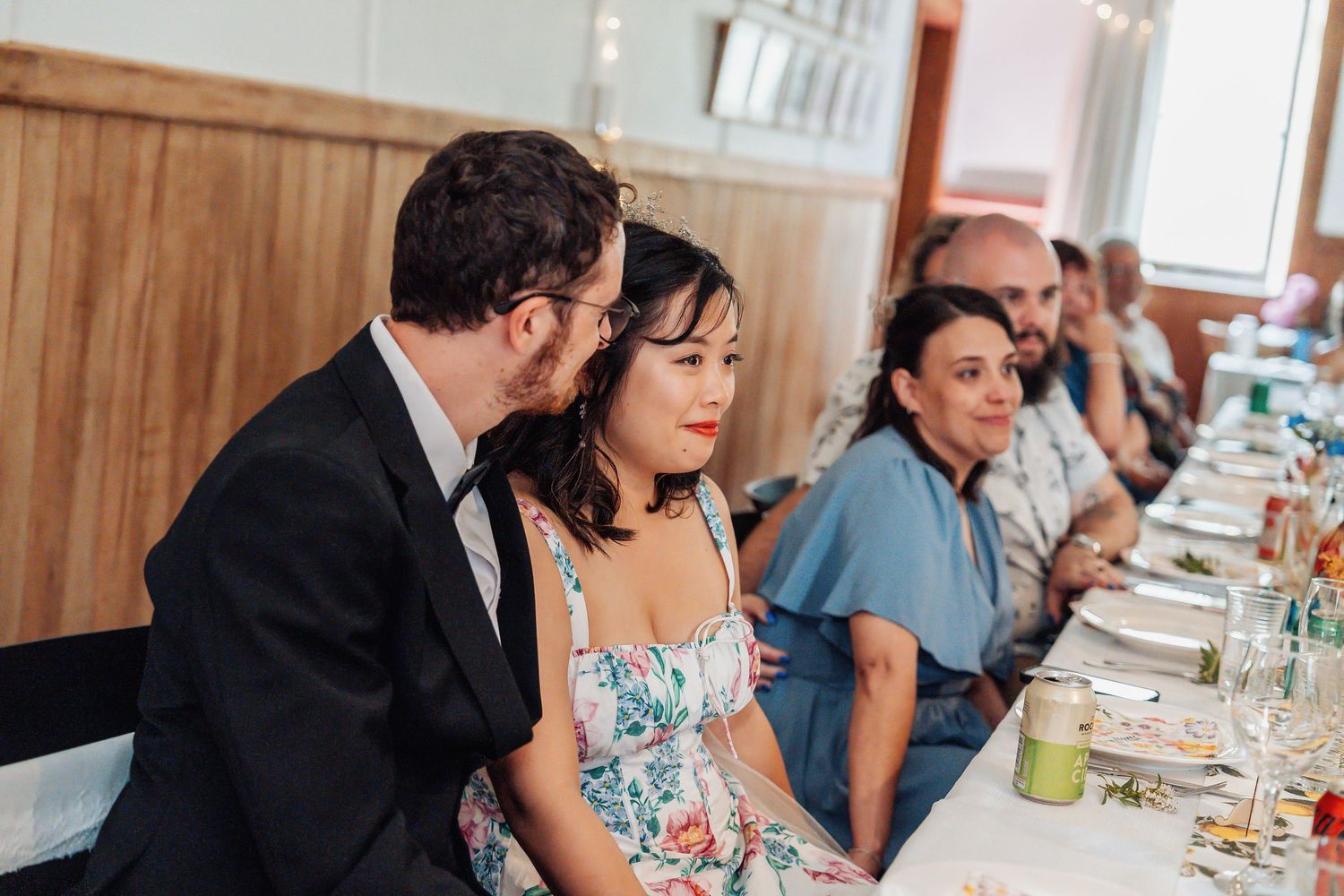 Groom whispering to bride in floral dress while seated at wedding reception dinner table.
