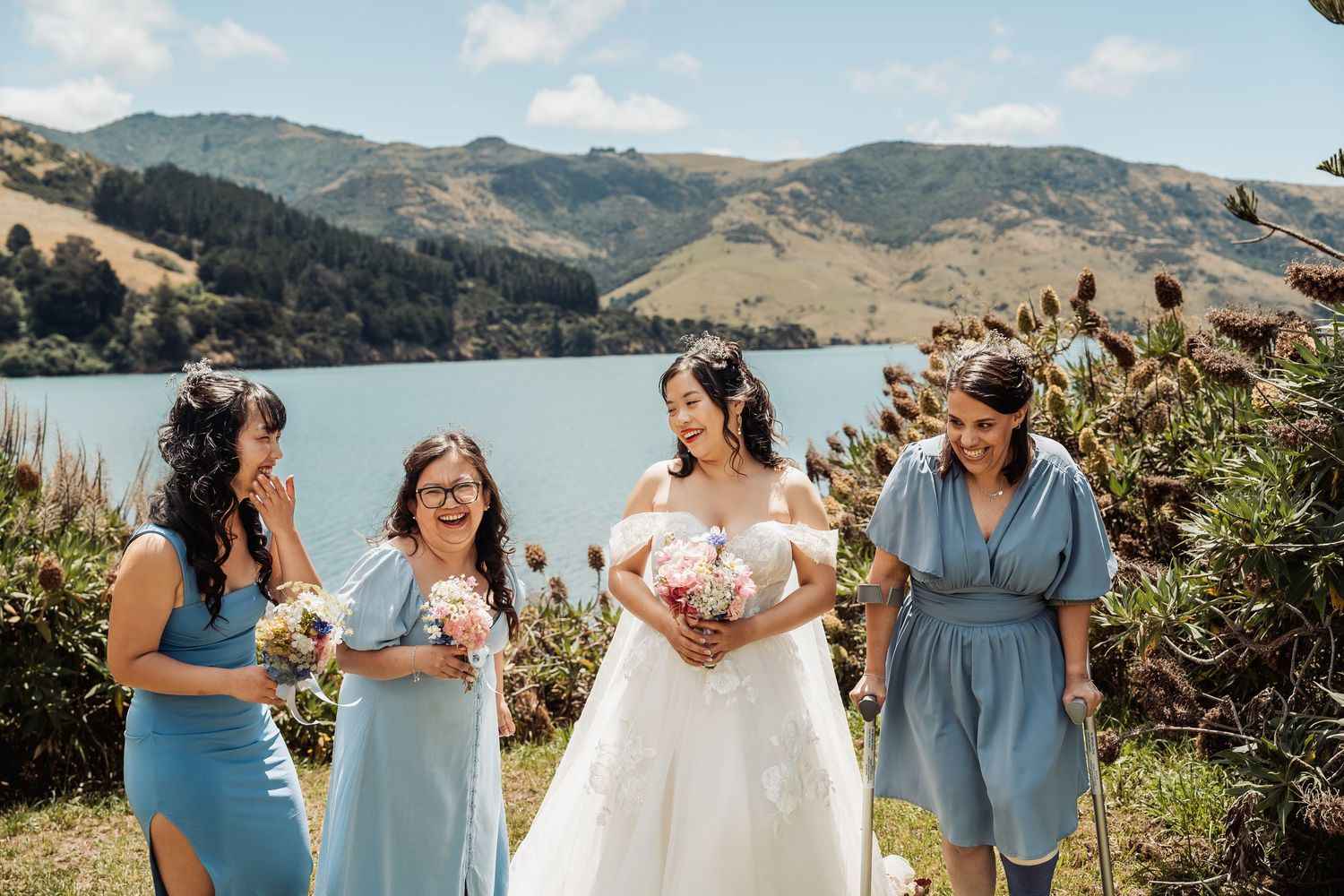 Bride in white gown with bridesmaids in blue dresses, holding bouquets by a scenic lake and mountains in New Zealand.