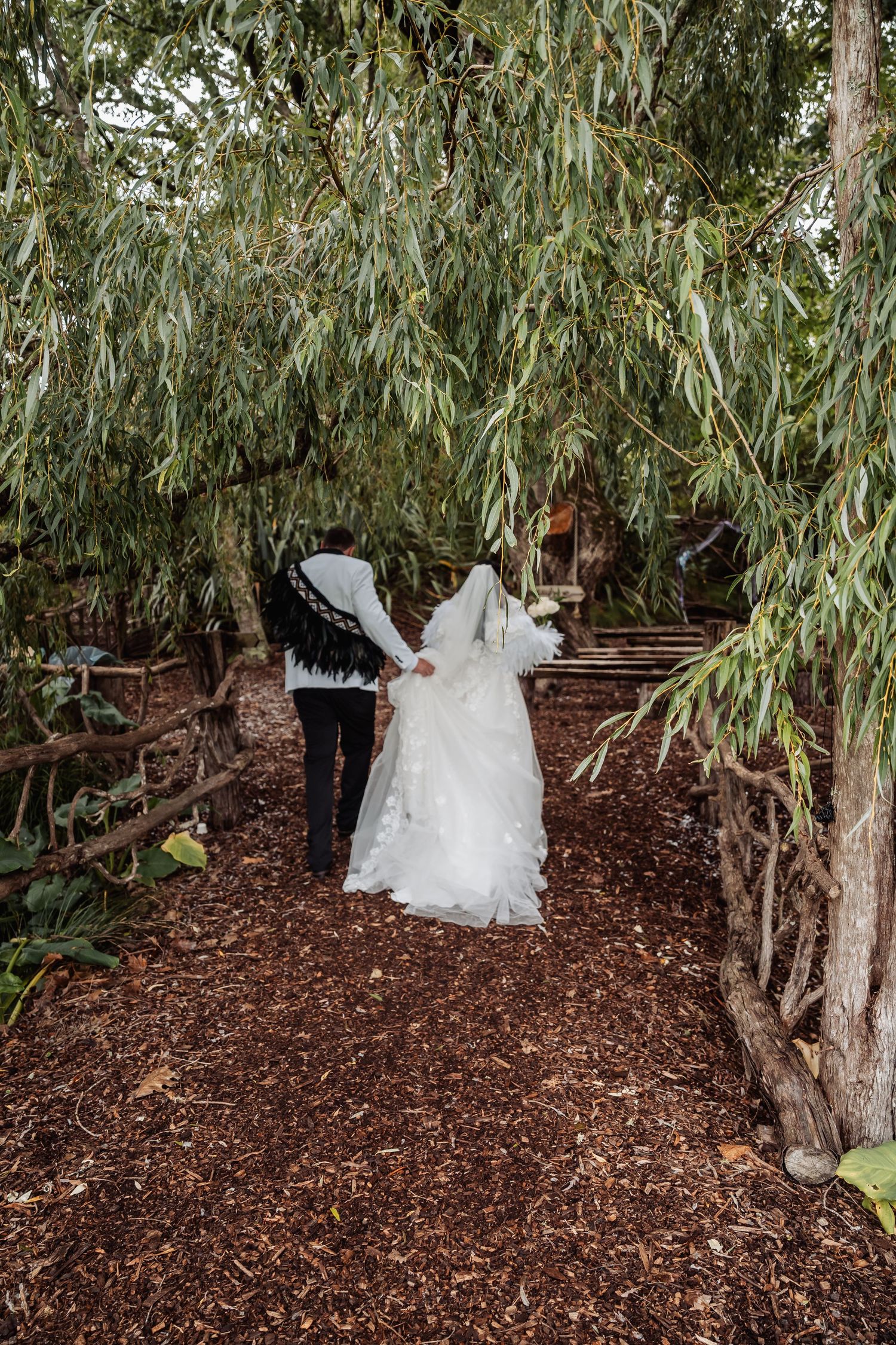 Bride and groom walk hand-in-hand along a mulch path under willow trees in a lush garden wedding setting.
