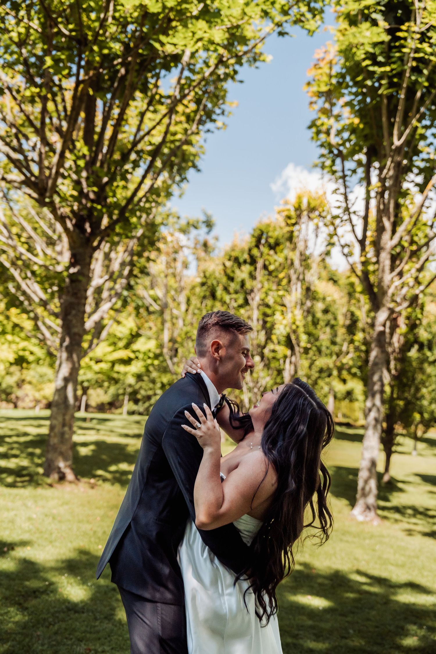 Bride and groom sharing a romantic kiss in a sunlit park with lush green trees in the background.