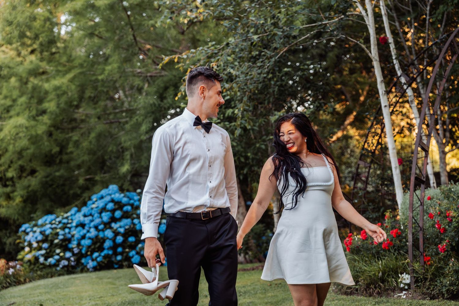 Happy newlyweds hold hands and laugh together while strolling through beautiful garden after wedding.