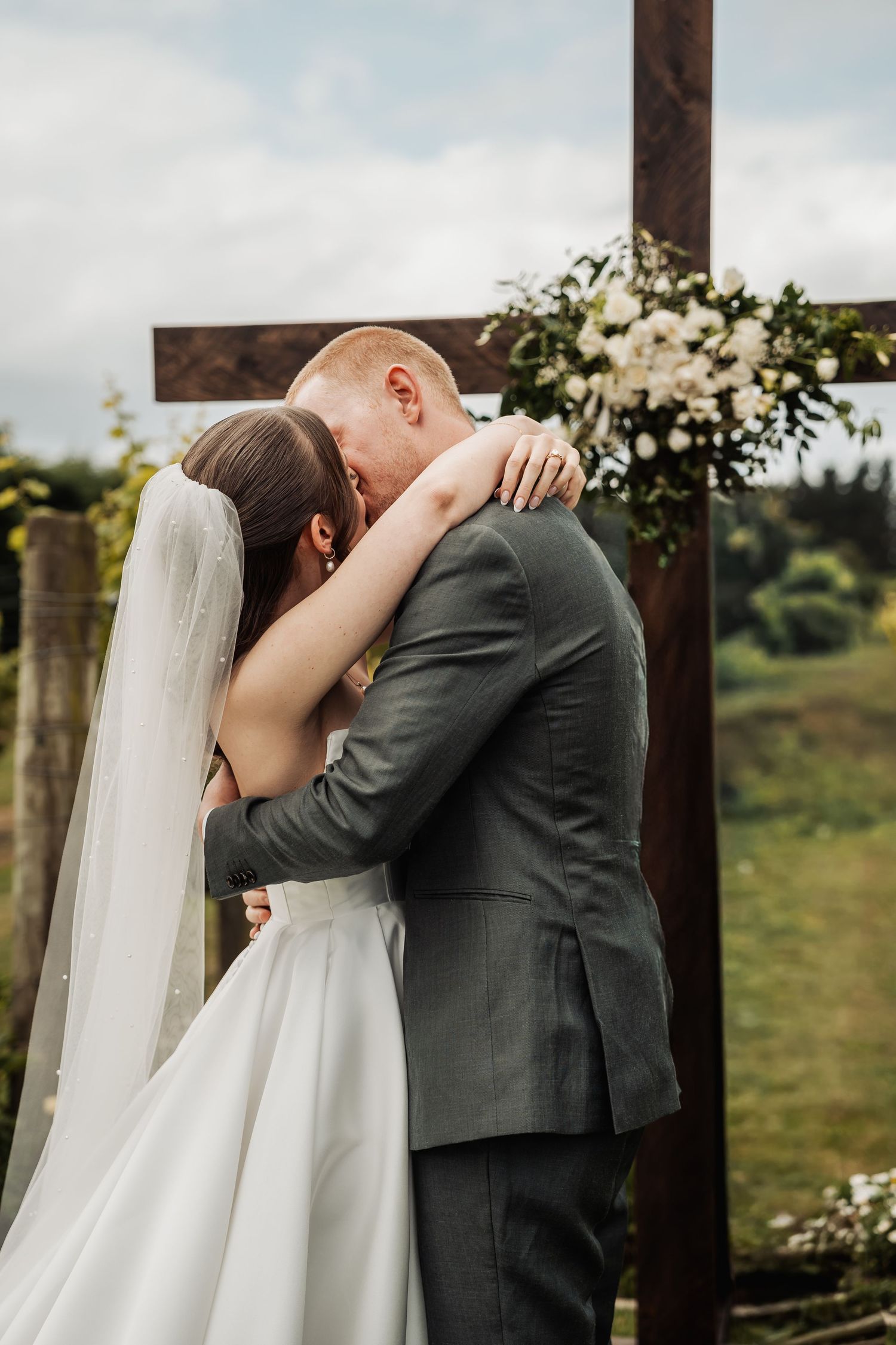 Newlyweds share their first kiss beneath a wooden cross altar adorned with white floral arrangements.