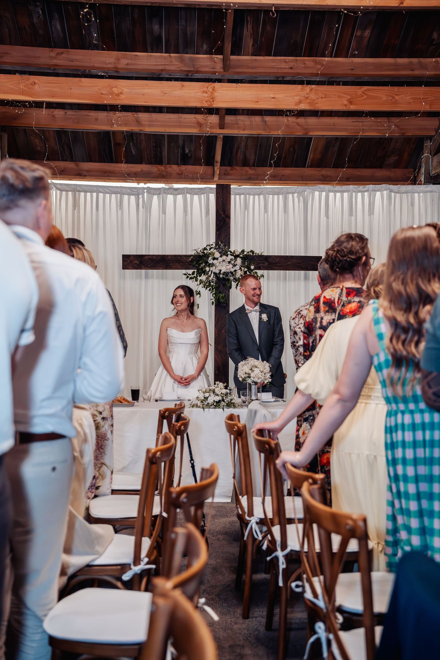 Bride and groom stand at altar during rustic barn wedding ceremony with guests seated in wooden chairs.