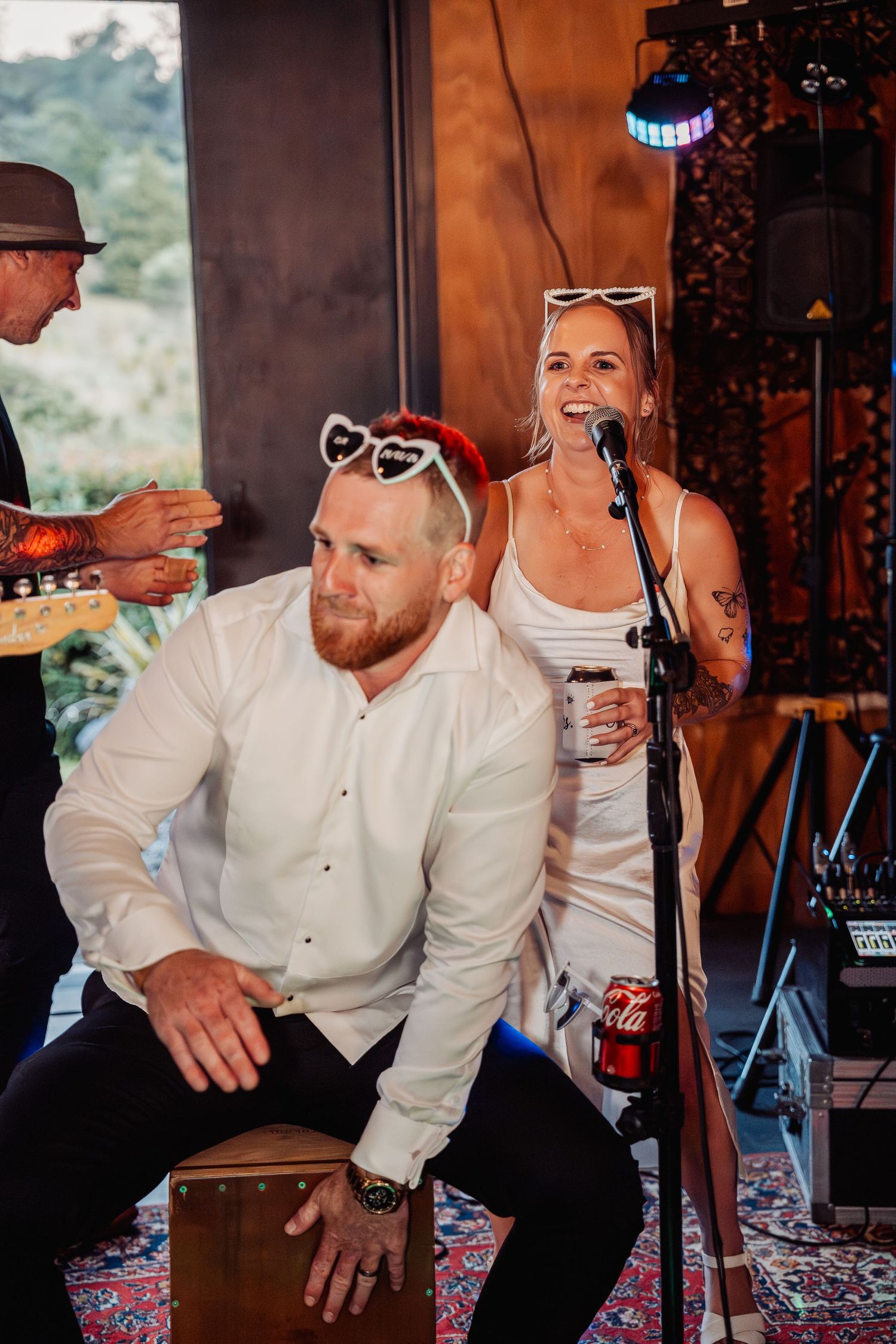 Groom in white shirt playing cajon drum beside laughing woman singing at wedding reception.