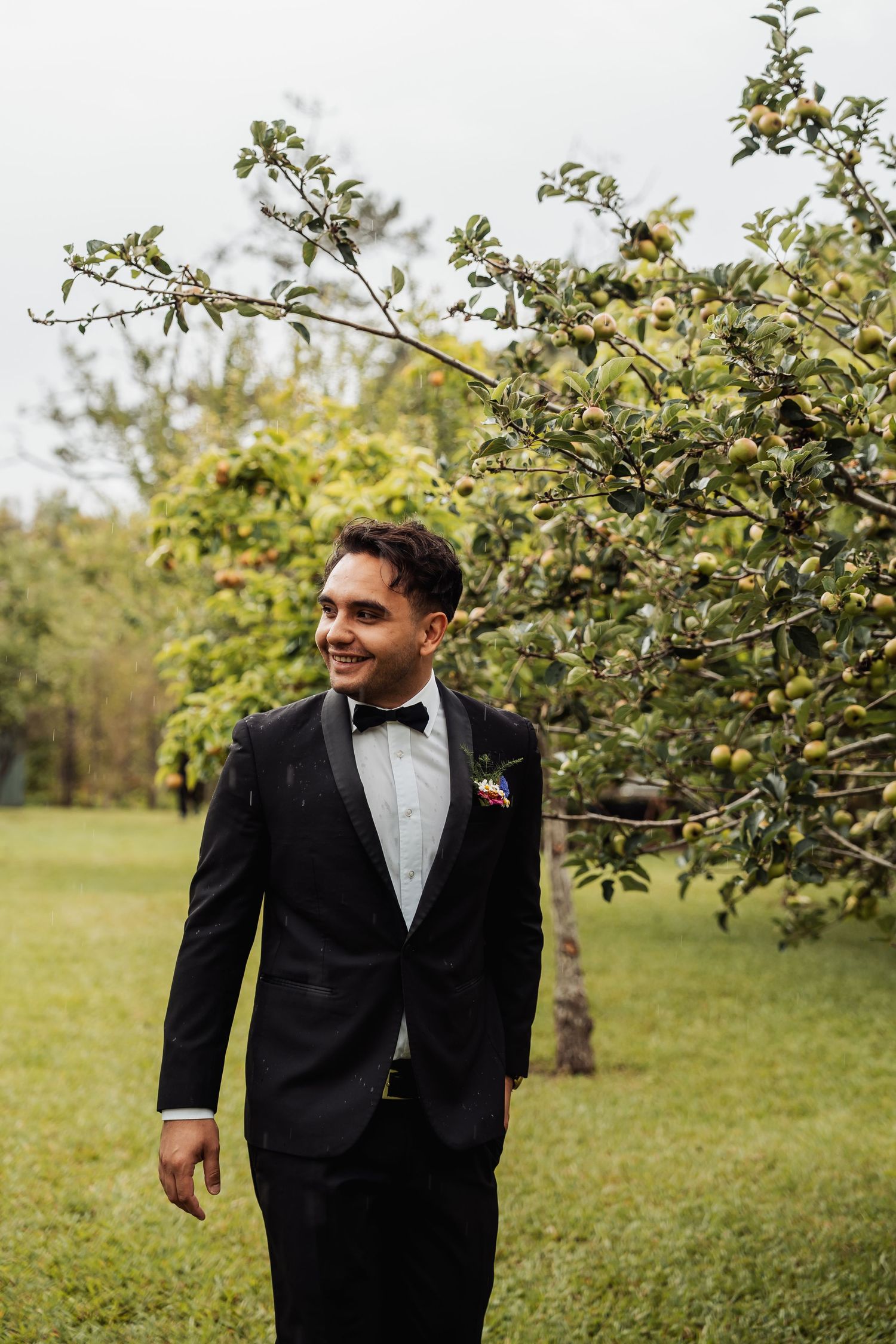 Smiling groom in black tuxedo with bow tie and boutonniere standing outdoors near apple tree.