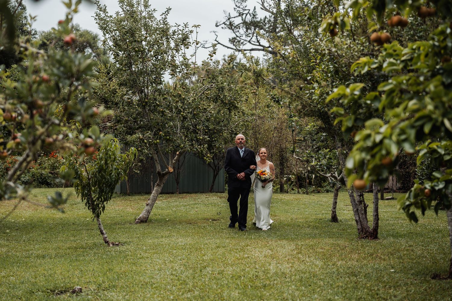 Bride and father walking through green orchard toward wedding ceremony.