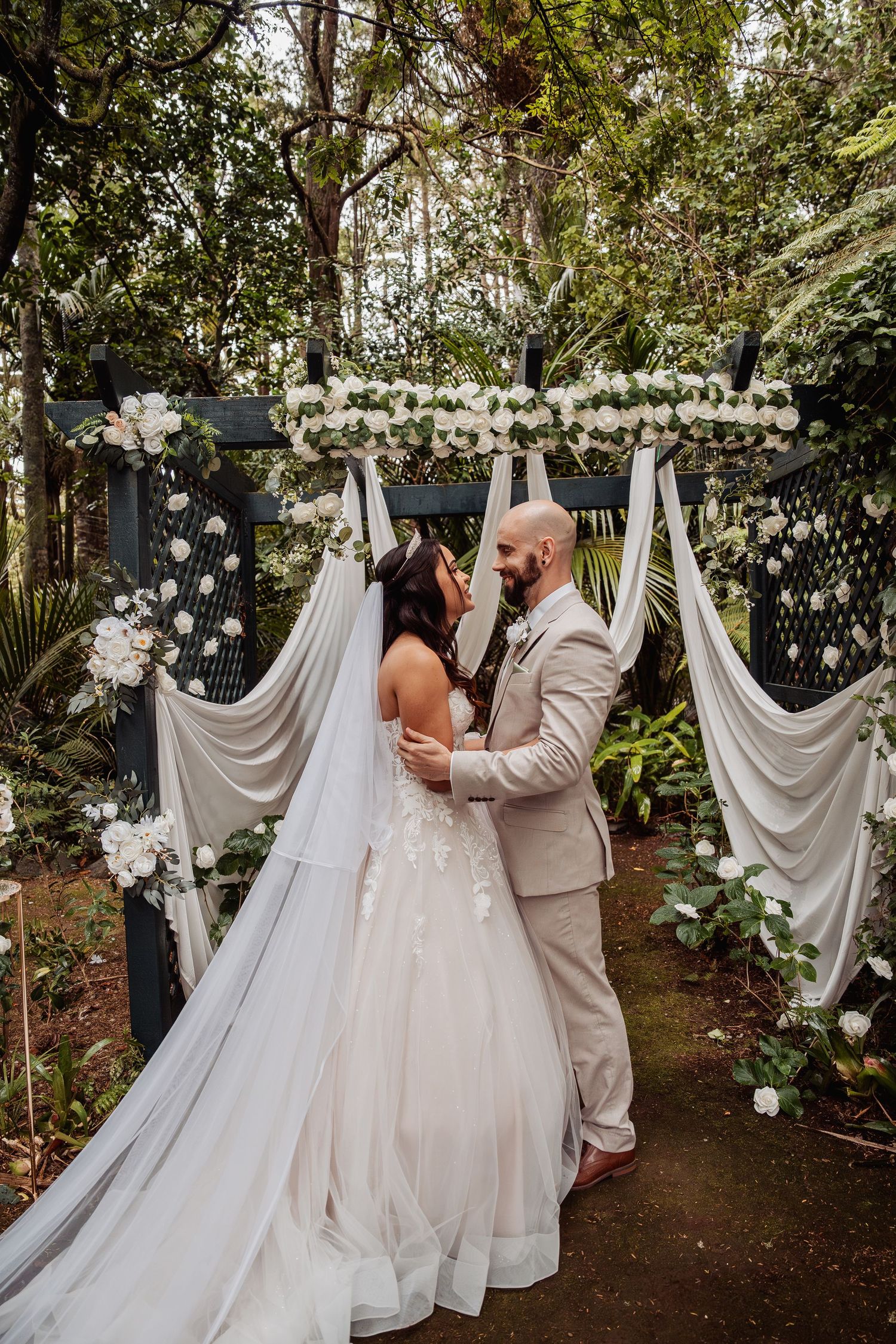 Bride and groom share a romantic moment at an outdoor floral wedding altar adorned with white roses and draped fabric.
