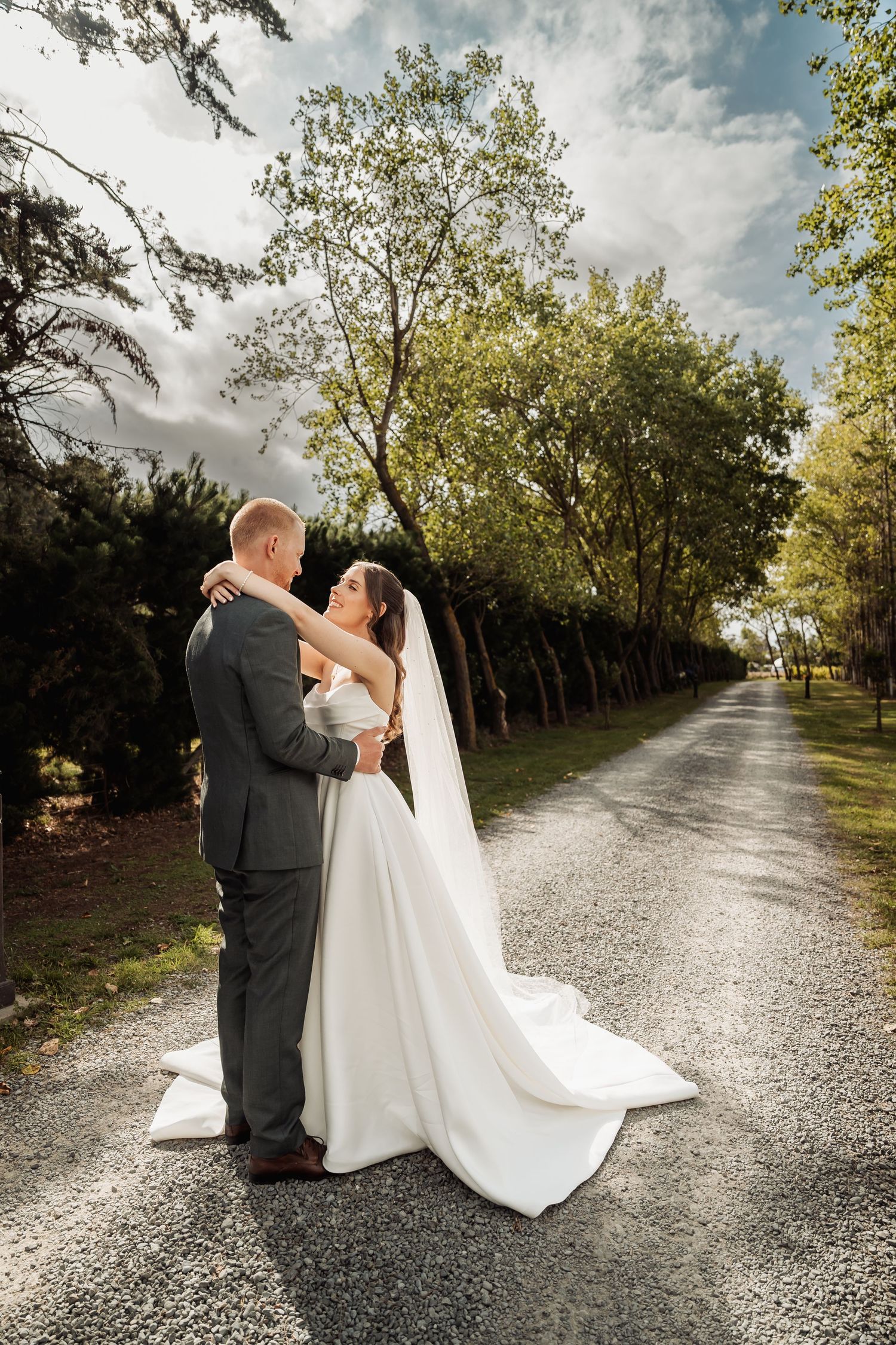 Bride and groom embrace on a tree-lined gravel path, bride in flowing white gown with veil, groom in gray suit.