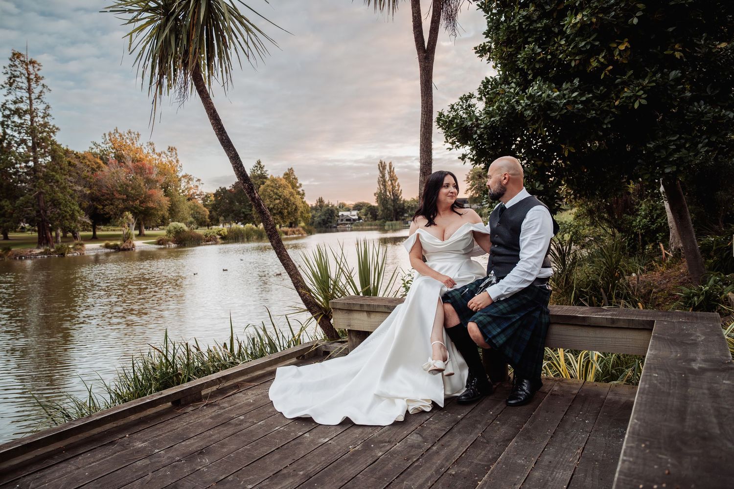 Bride and groom sitting together on a wooden deck beside a tranquil lake at sunset, groom wearing a tartan kilt.