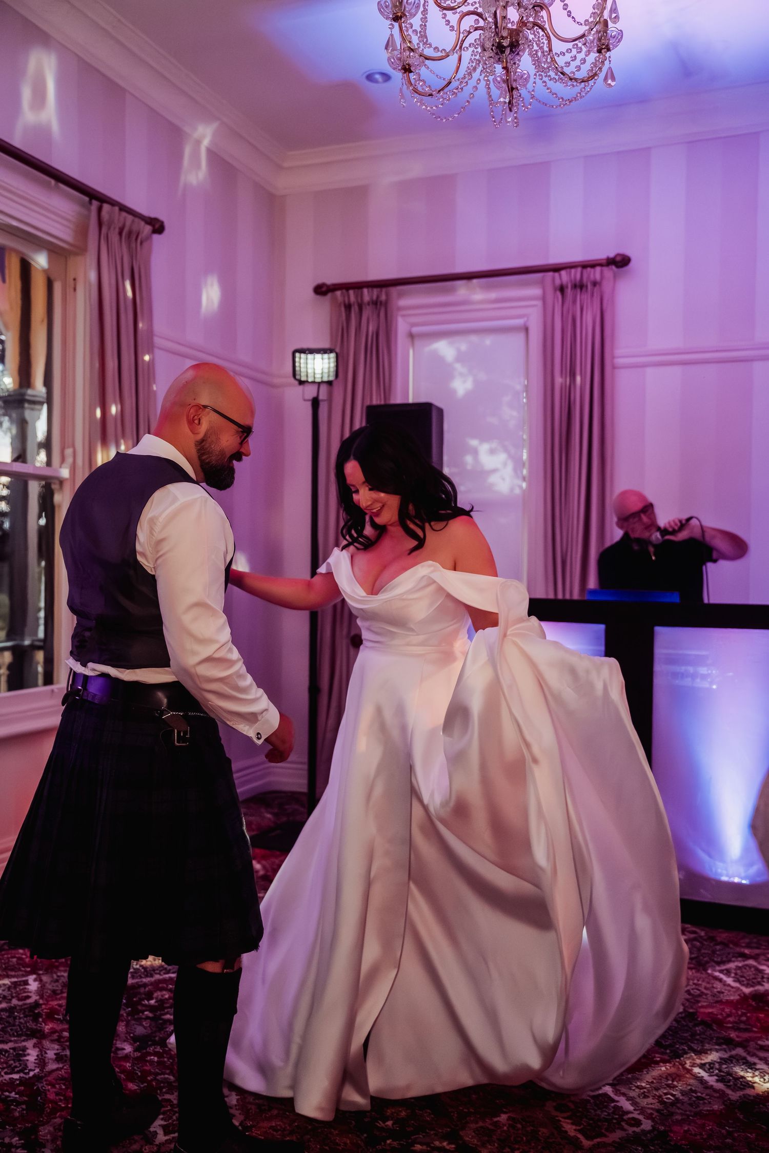 Bride in white gown and kilted groom dance at elegant chandelier-lit wedding reception.
