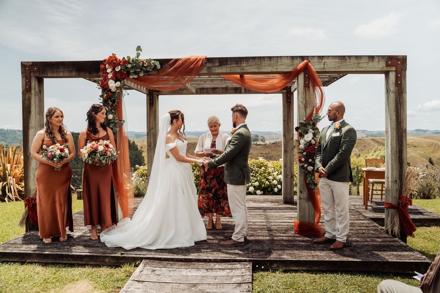 Wedding ceremony under rustic wooden arch with bridesmaids, groomsmen, and rolling hills behind.