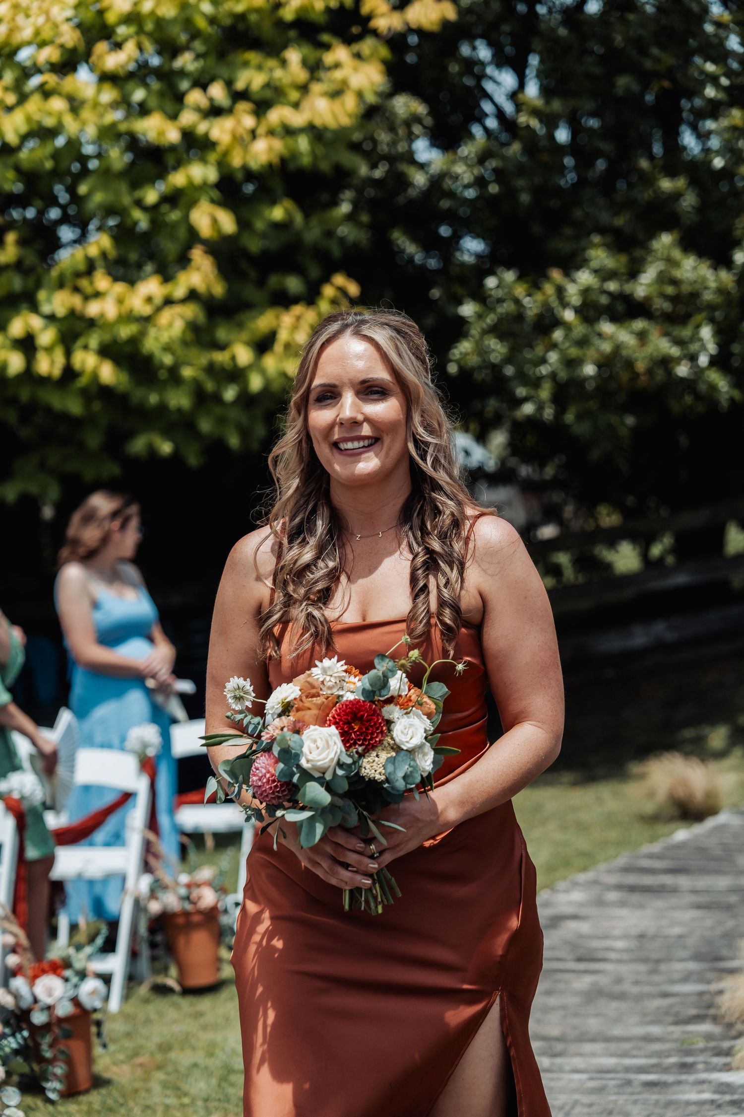 Smiling bridesmaid in rust orange dress walking down aisle holding colorful bouquet at outdoor wedding.