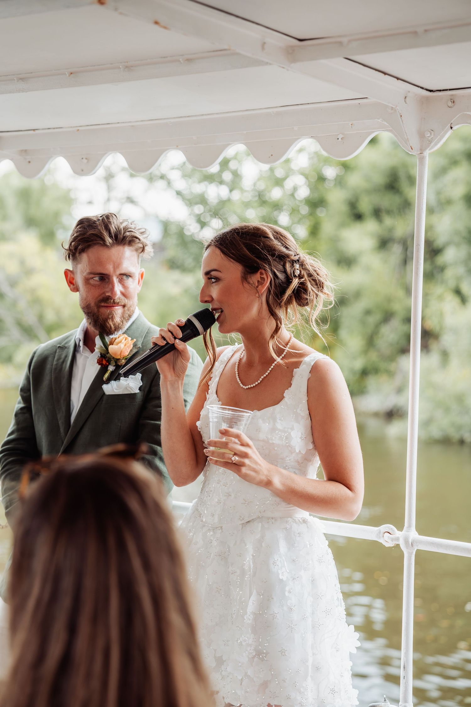 Bride speaking into microphone beside groom in green suit during outdoor waterside wedding reception.