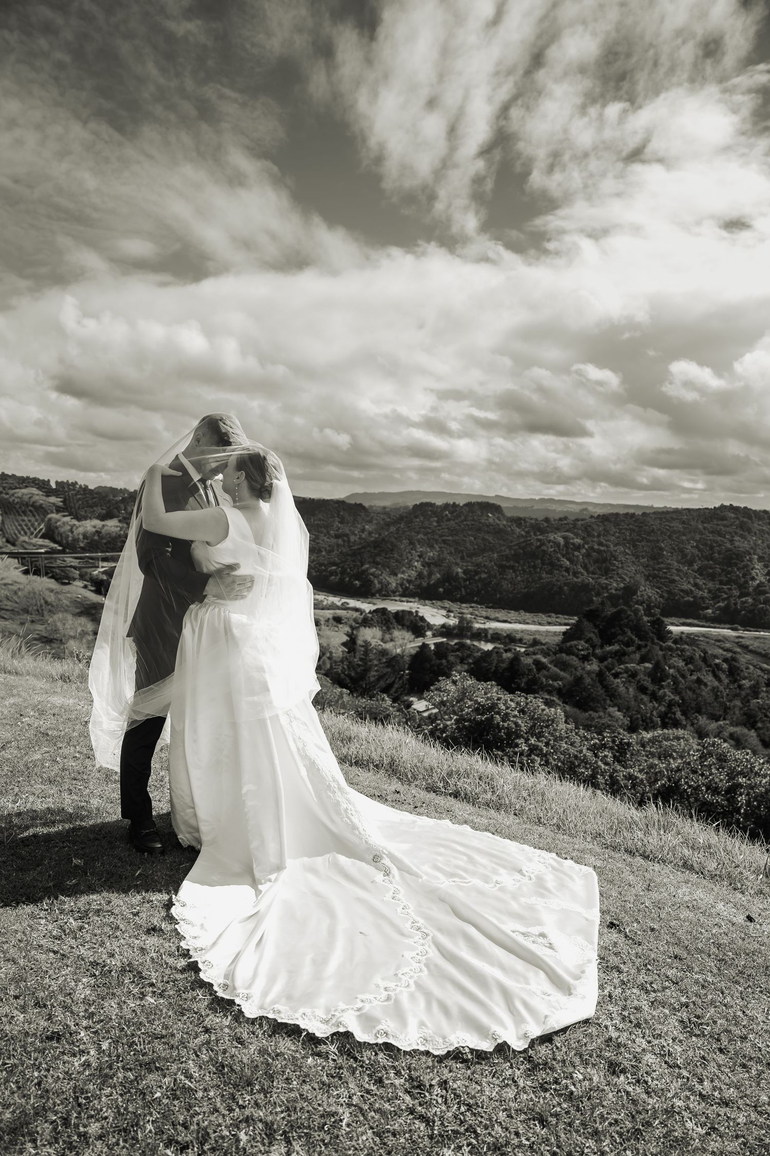Black and white photo of a bride and groom embracing on a hilltop with a scenic valley and cloudy sky backdrop.