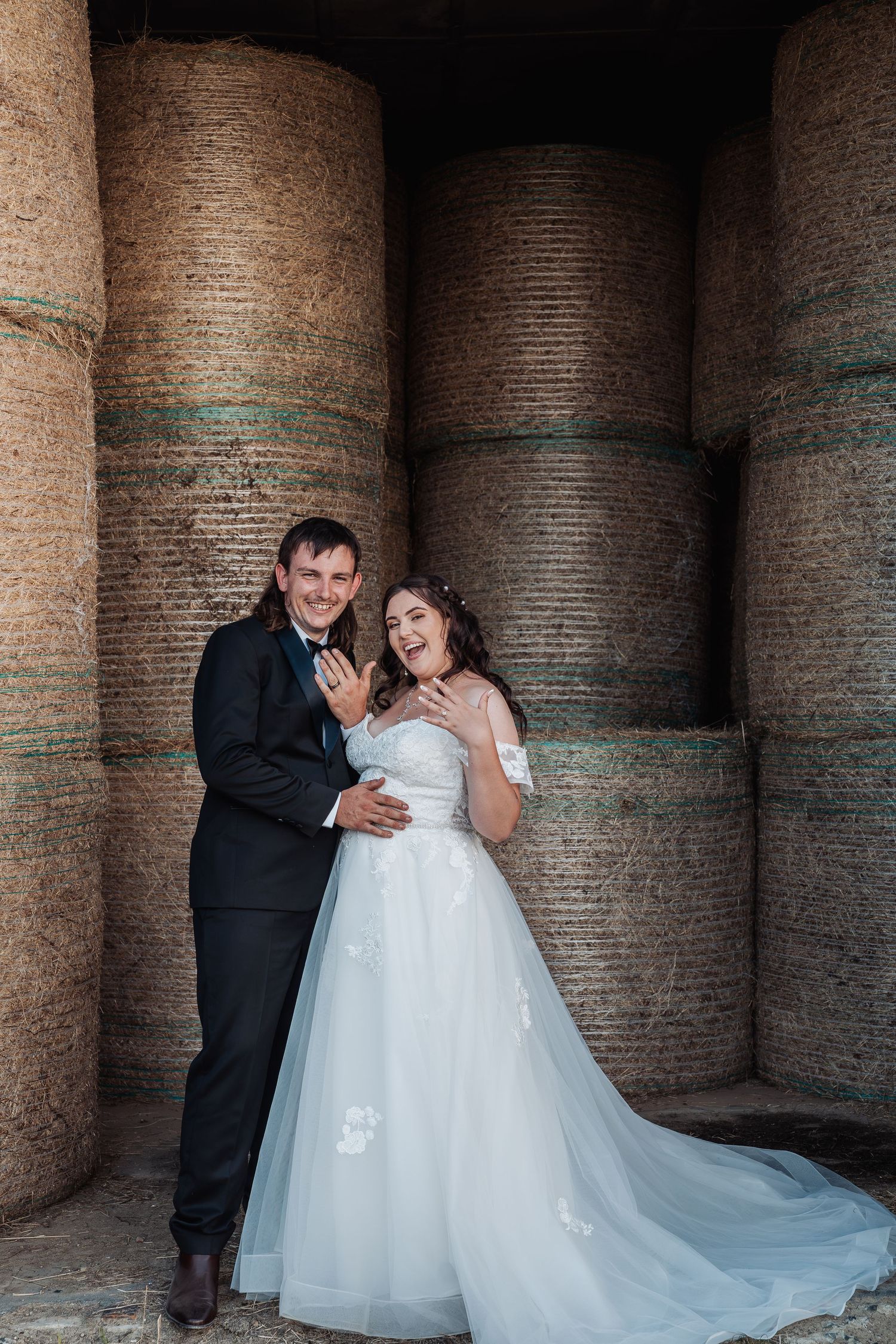 Laughing bride and groom pose together in front of large round hay bales in rustic barn setting.