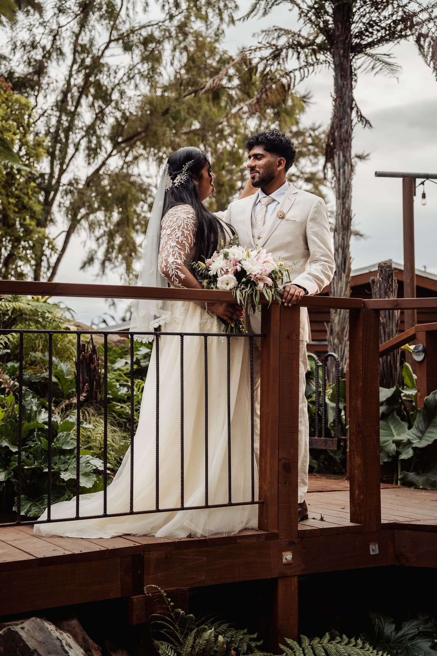 Bride and groom in white wedding attire stand on a wooden deck surrounded by lush tropical greenery.