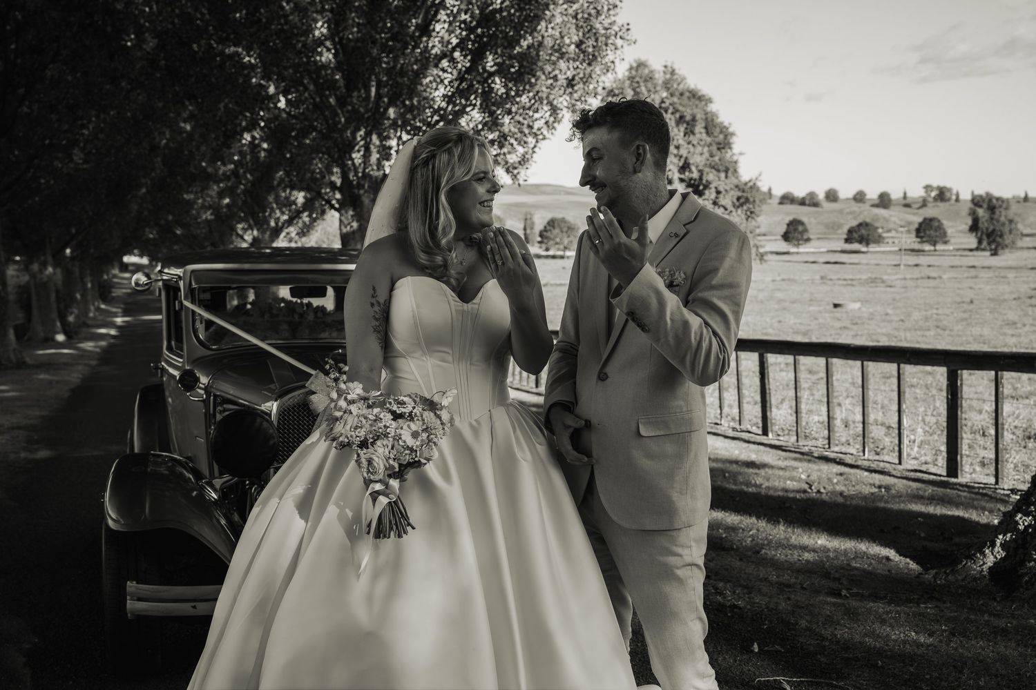 Black and white photo of bride and groom sharing a tender moment beside a vintage car outdoors.