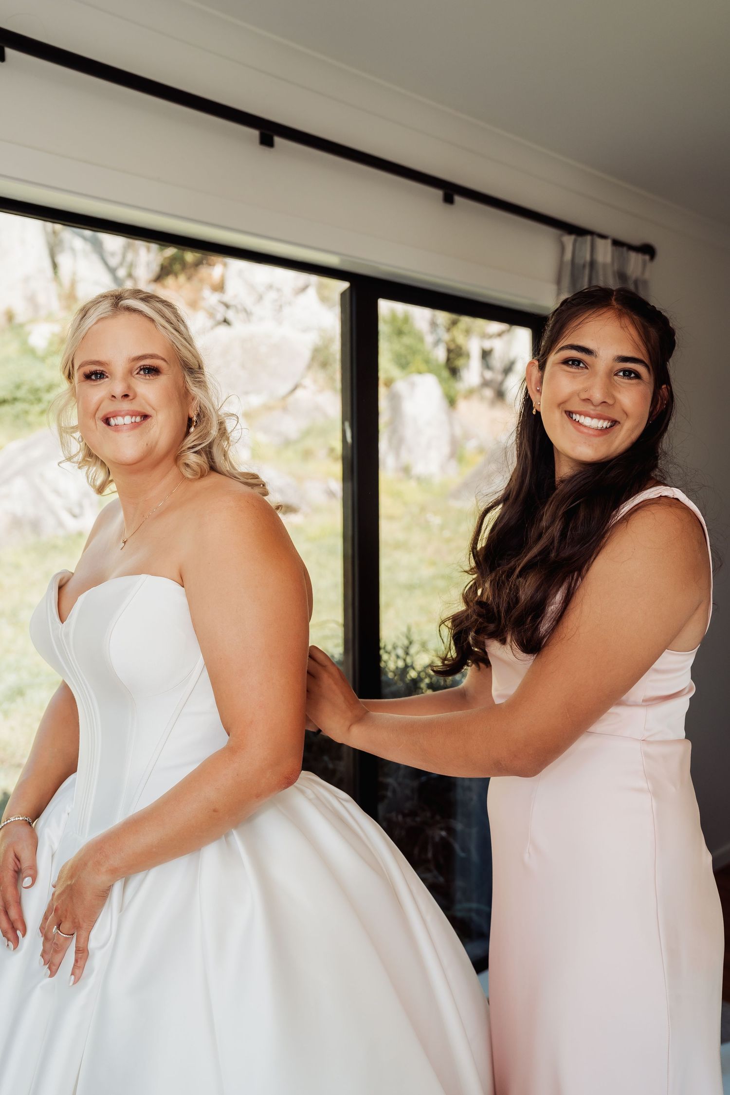 A bridesmaid in blush pink helps zip up a bride in a strapless white ballgown wedding dress.